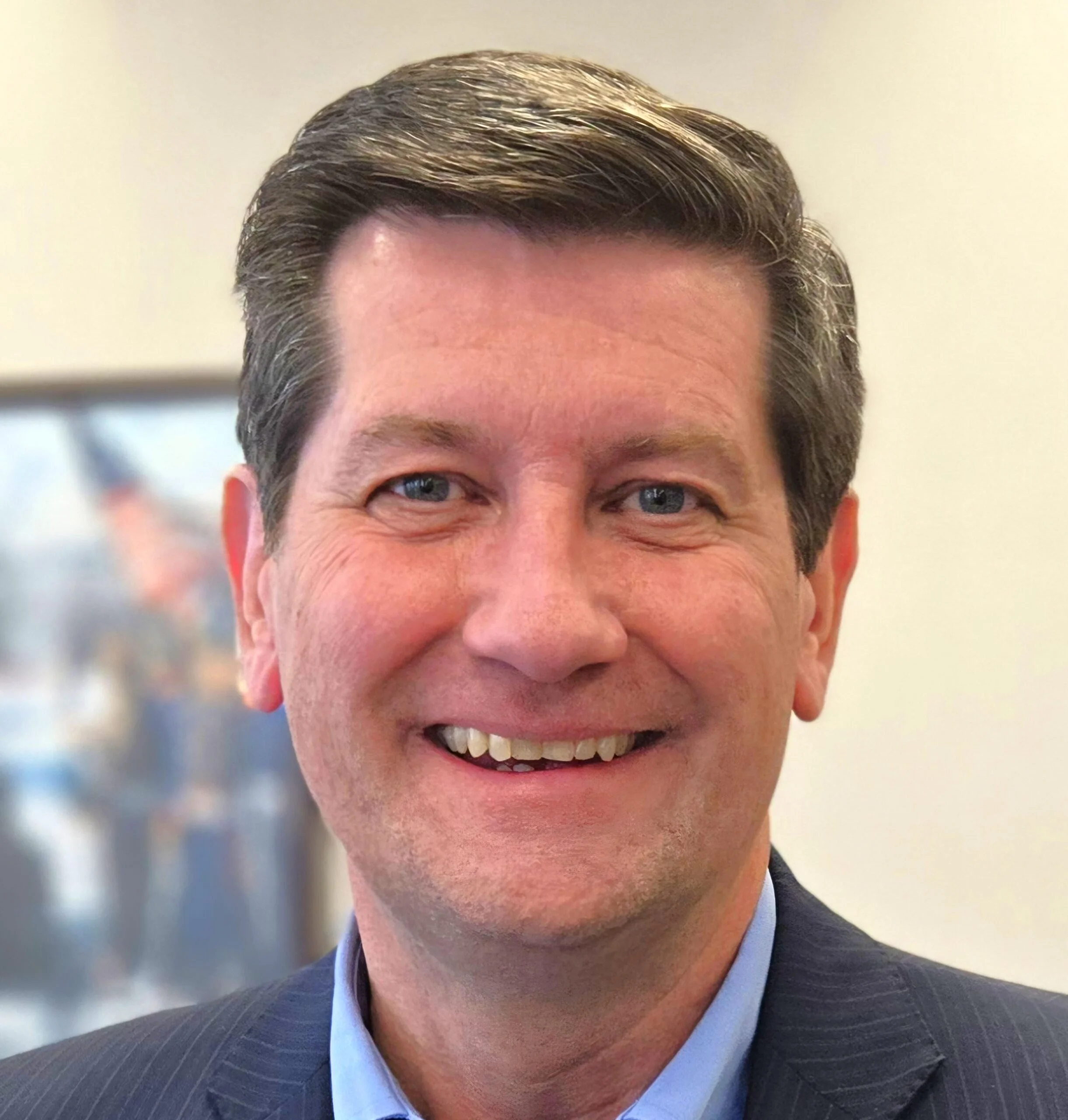 Close-up portrait of a middle-aged man with dark hair and blue eyes, smiling, wearing a navy suit and light blue shirt.