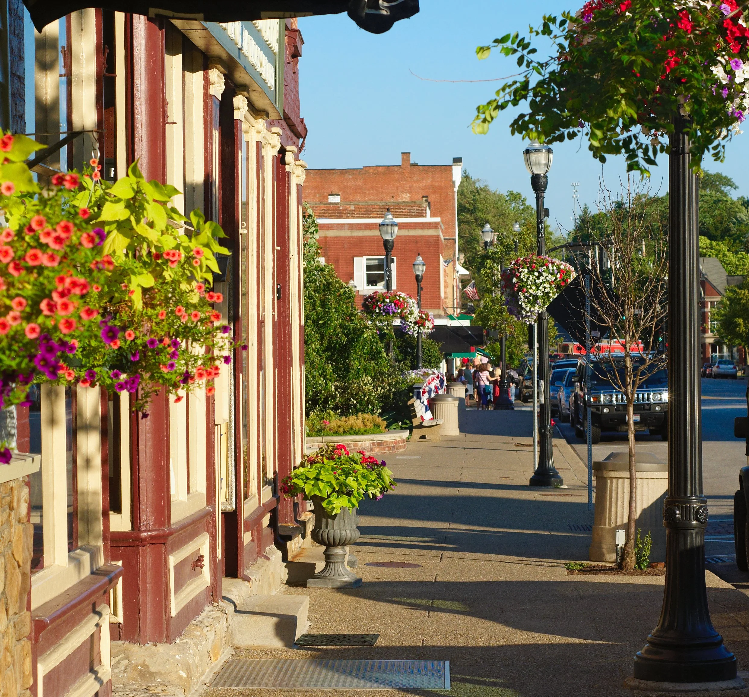A charming downtown street lined with flowers, vintage lampposts, and brick buildings, with people walking and parked cars in the background.