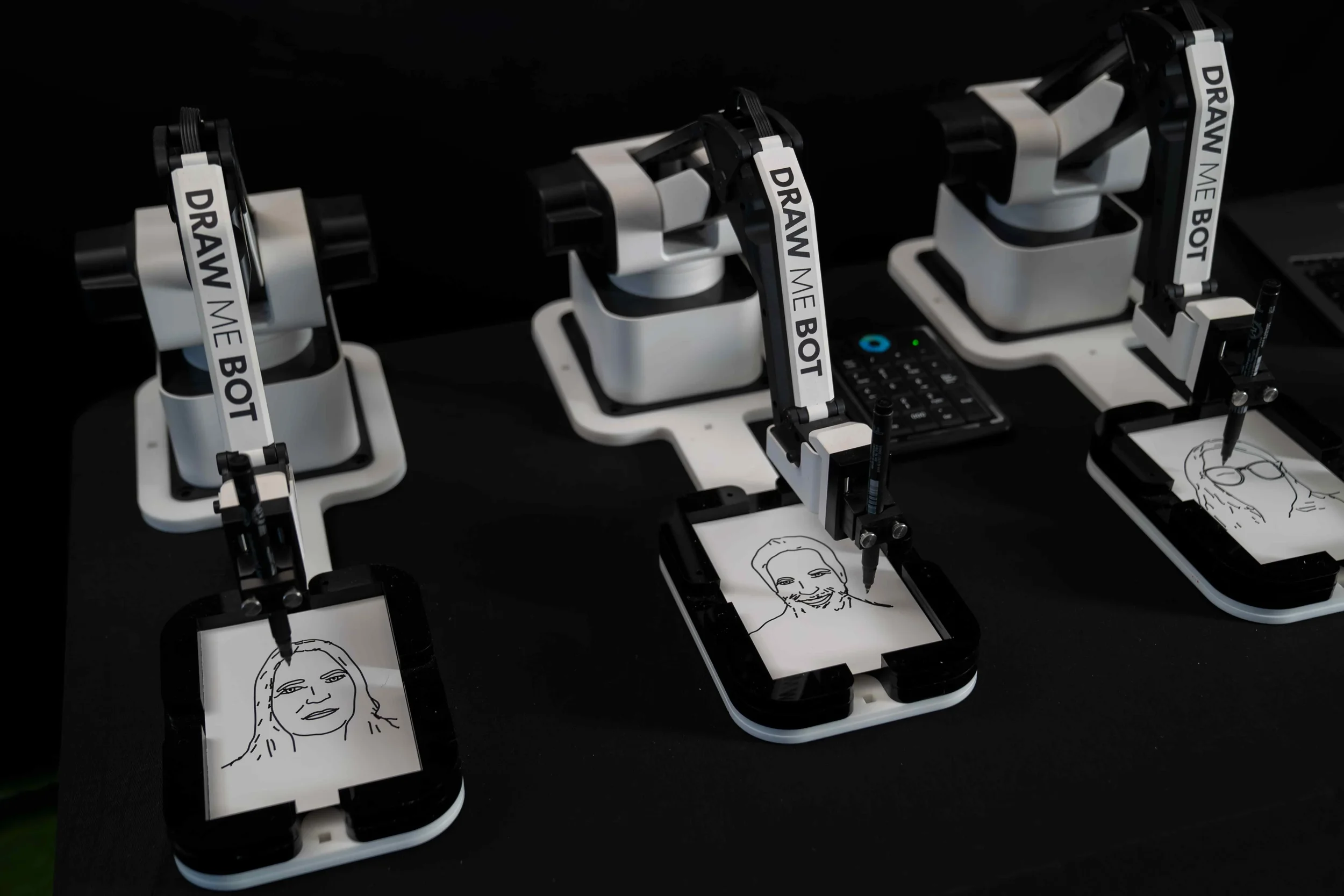 Three robotic drawing machines each creating a portrait sketch on a mobile device with the phrase 'Draw me bot' on their arms.