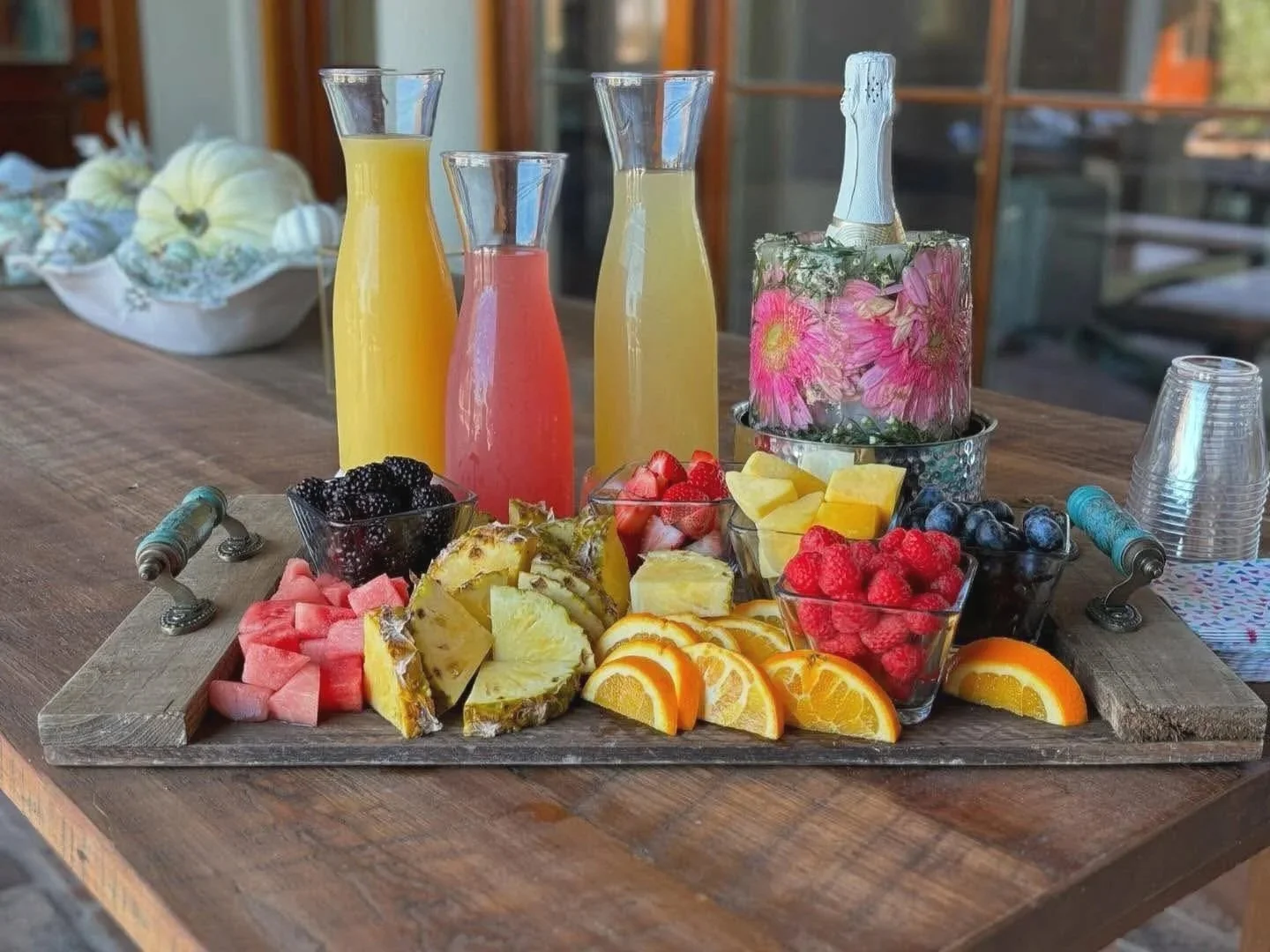 A wooden table set with glasses of juice, bowls of mixed berries and sliced fruit, and a bottle of sparkling wine with a floral wrapped bottle holder.