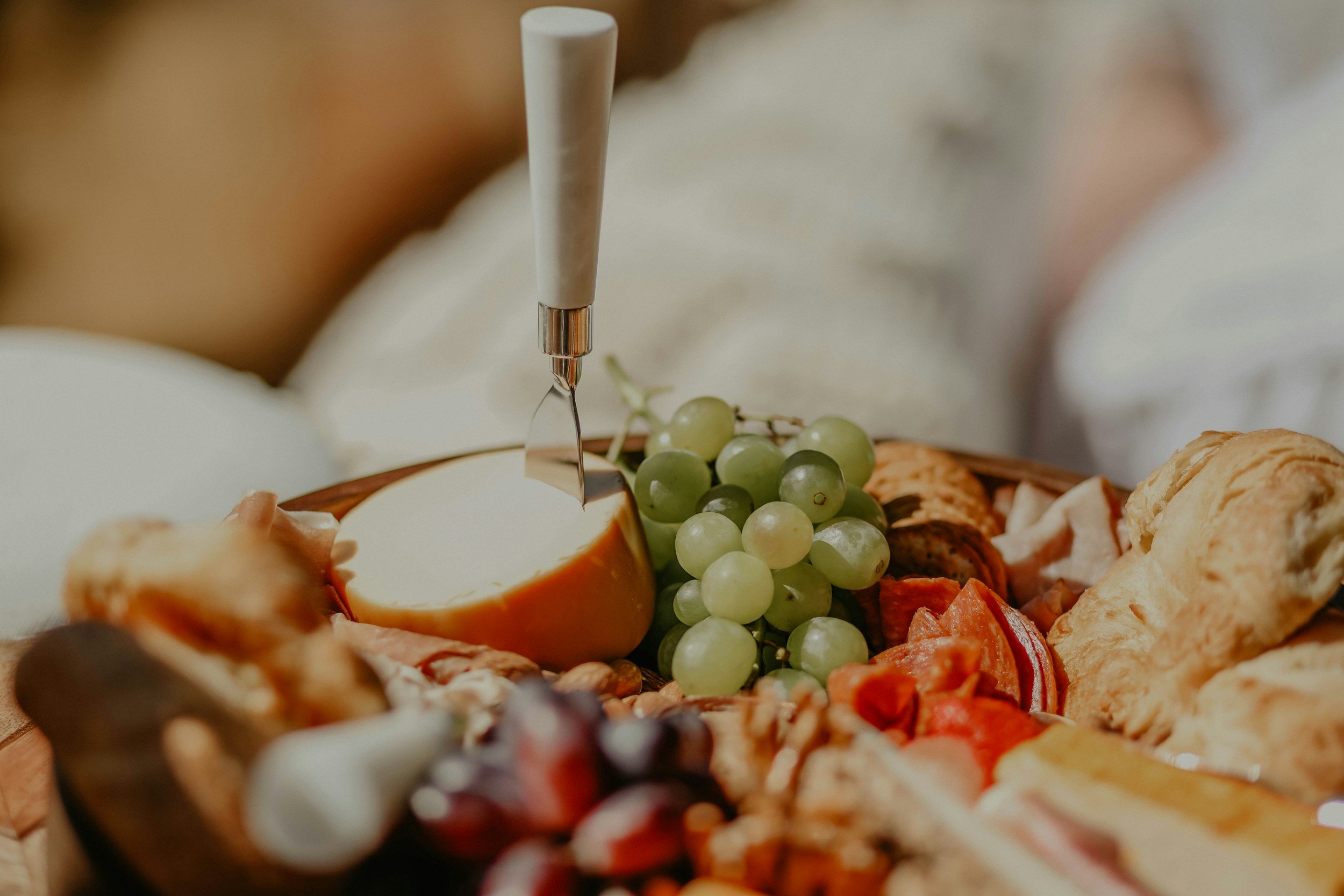 A cheese knife cutting into a wheel of cheese on a cheese board with grapes, crackers, and cured meats.