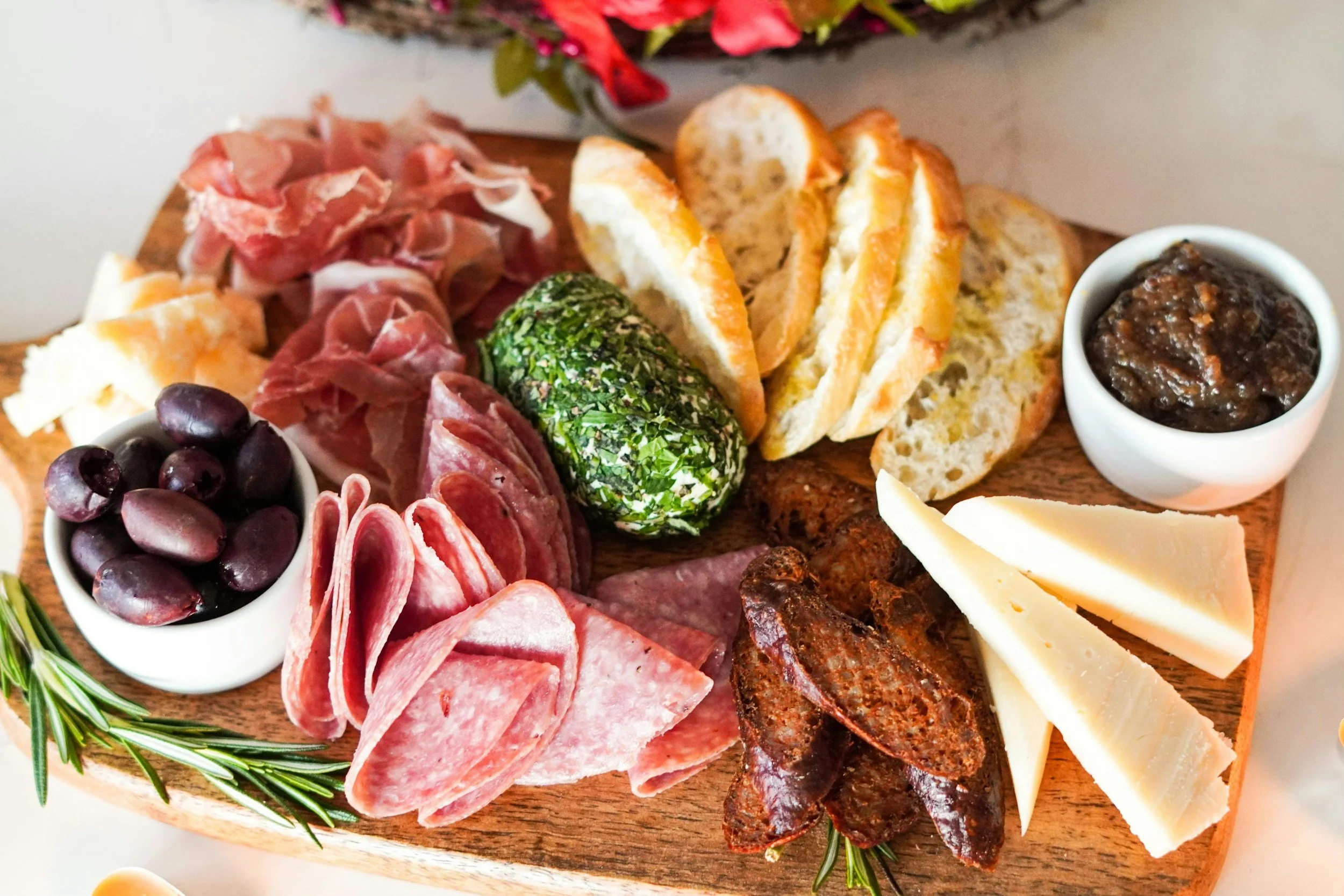 A wooden charcuterie board with an assortment of cheese, cured meats, bread slices, olives, fig jam, and garnished with rosemary.