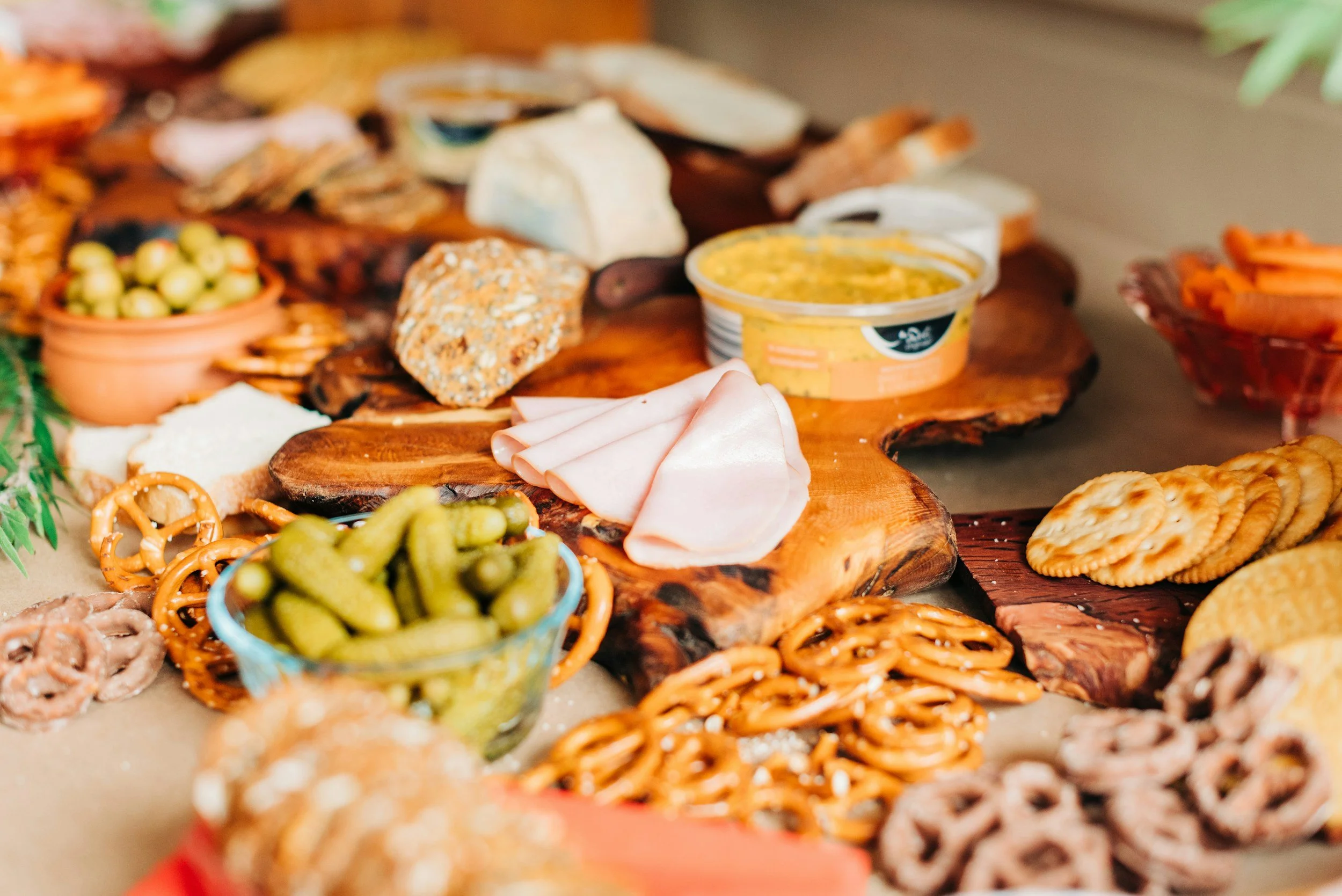 A charcuterie board with an assortment of cheeses, crackers, pickles, sliced meats, hummus, and various snacks arranged on a wooden board and surrounding platters.