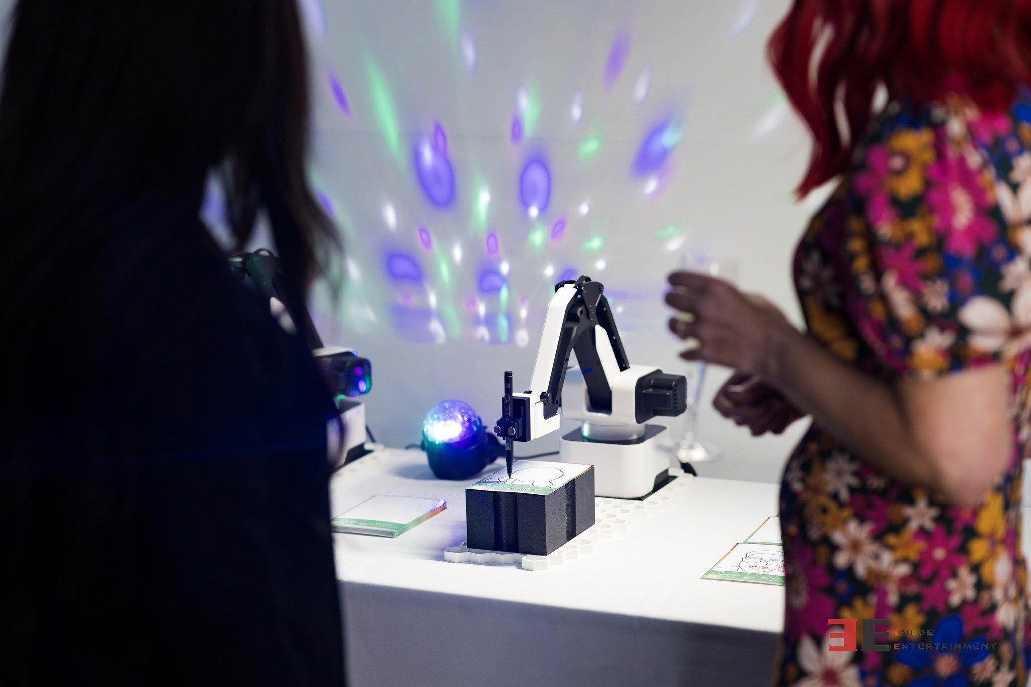 A robotic arm is conducting a drawing activity on a piece of paper, with colorful laser lights projected on the wall behind it. Two women are standing nearby, observing the activity.