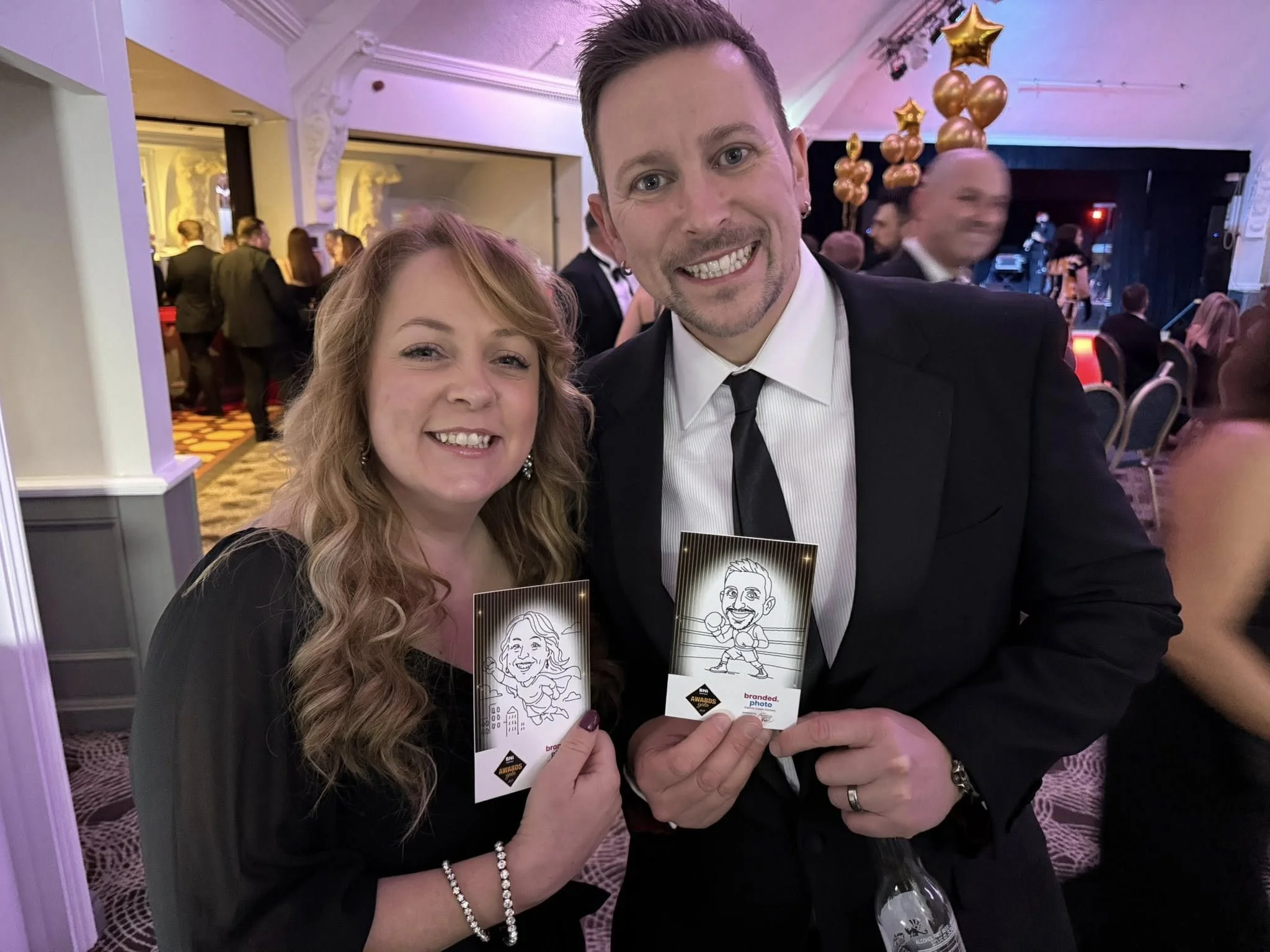 A woman and a man in formal attire smiling and holding promotional cards at a party or awards event, with a live stage and balloon decorations in the background.