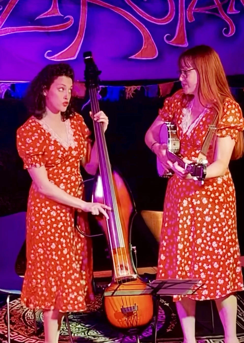 Two women in matching red floral dresses performing with musical instruments on stage. One is playing a double bass, and the other is playing a ukulele with a microphone stand in front of her. The background has purple and pink lighting.