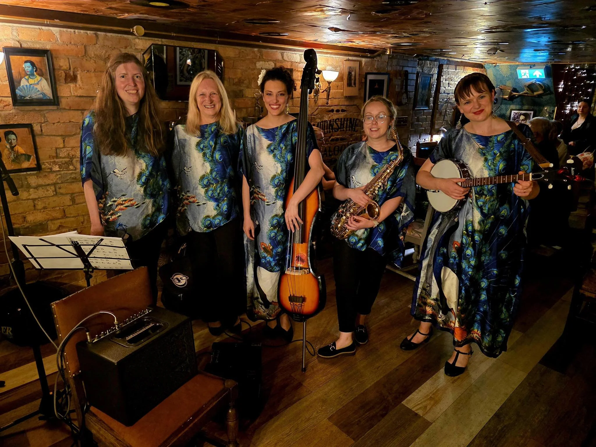 Five women standing together in a cozy music venue, holding musical instruments including a double bass, saxophone, and banjo, all wearing matching colorful top with a peacock feather design, with framed art on the brick wall behind them and patrons seated in the background.
