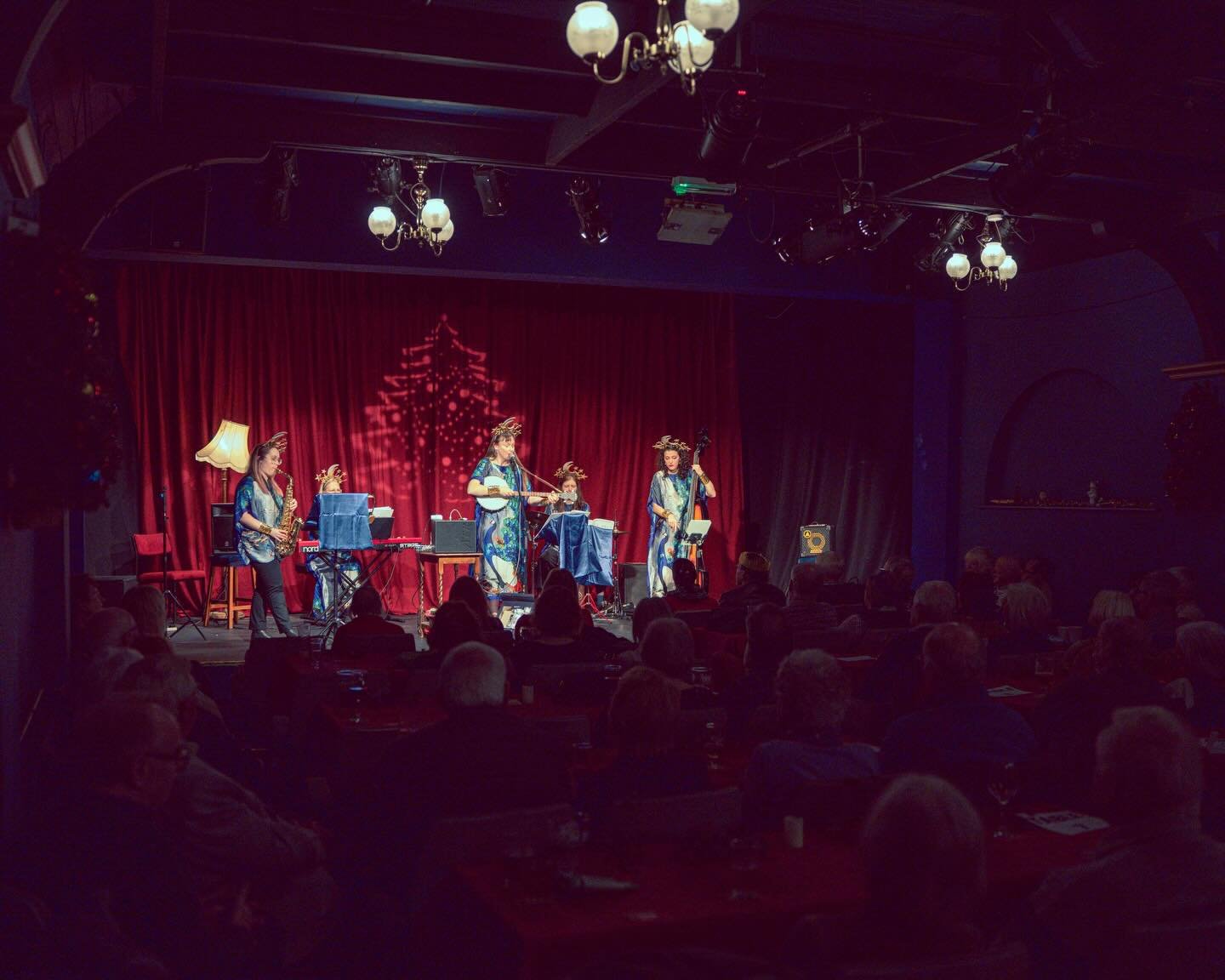 A group of female musicians performing on a stage with a red curtain and Christmas tree shadow projection, decorated with reindeer antler headbands, in front of an audience in a dimly lit venue.