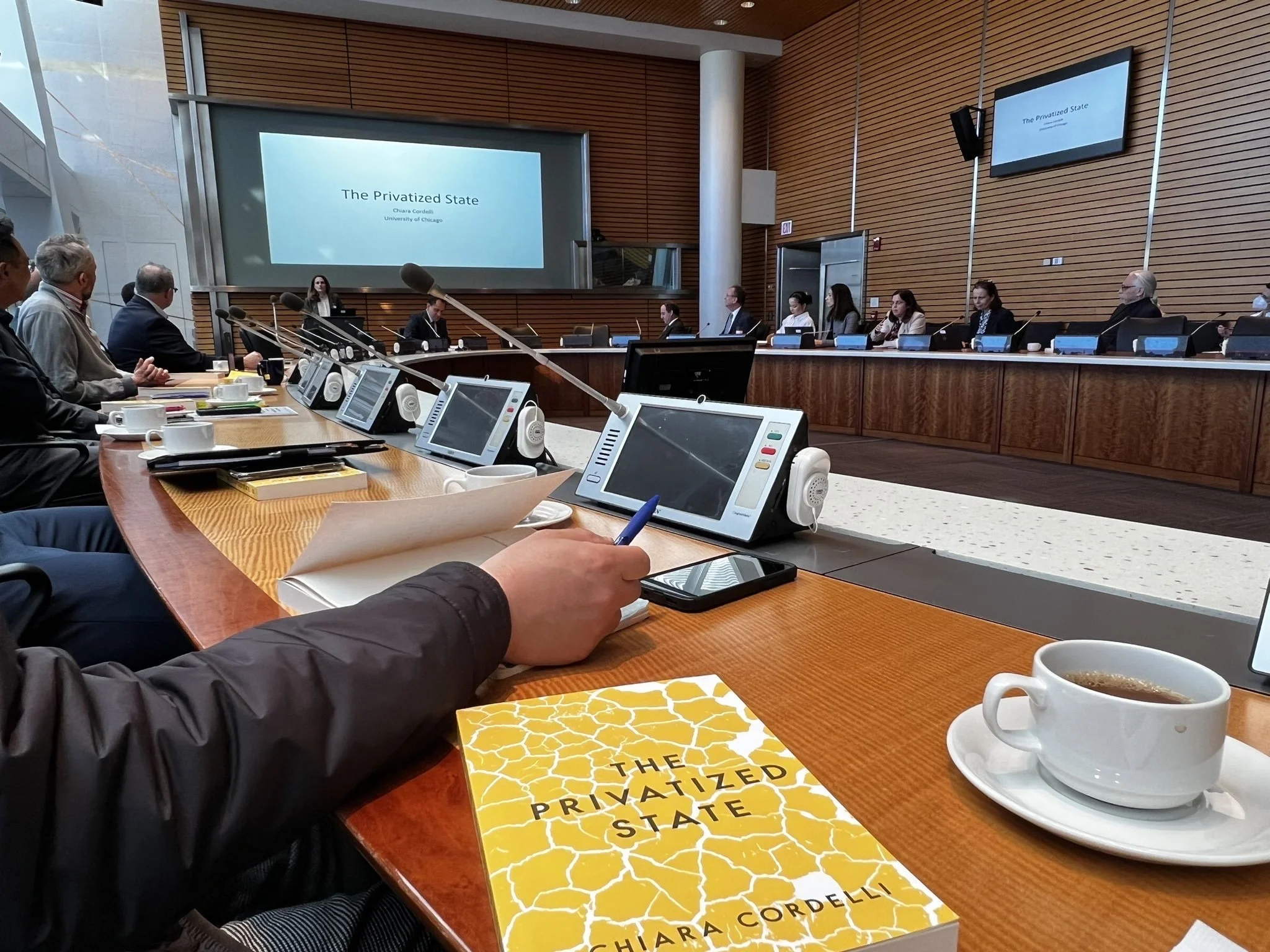 A conference room with a long table and multiple speakers and attendees. There are monitors on the table, some with microphones, and a presentation screen at the front displaying the title 'The Privatized State' by Chiara Cordelli. In the foreground, a person is taking notes with a pen, a cup of coffee, and a book titled 'The Privatized State' by Chiara Cordelli.