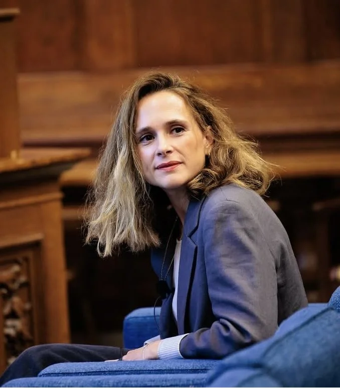 A woman with wavy blonde hair wearing a gray blazer, sitting on a blue chair in a wooden-paneled room.