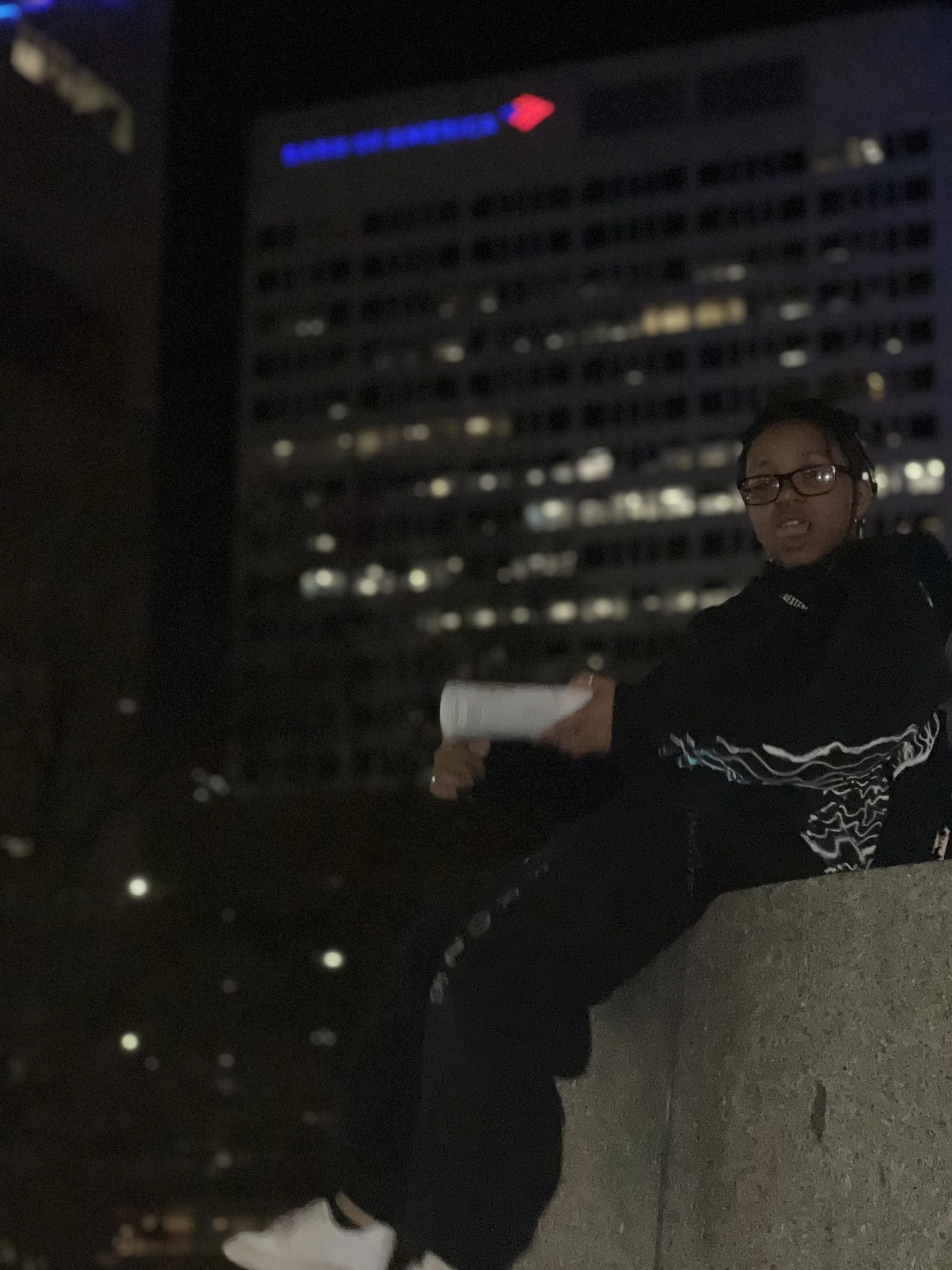 A person wearing glasses and dark clothing sitting outside at night, holding a book or notepad, with a cityscape and a tall building with illuminated windows in the background.