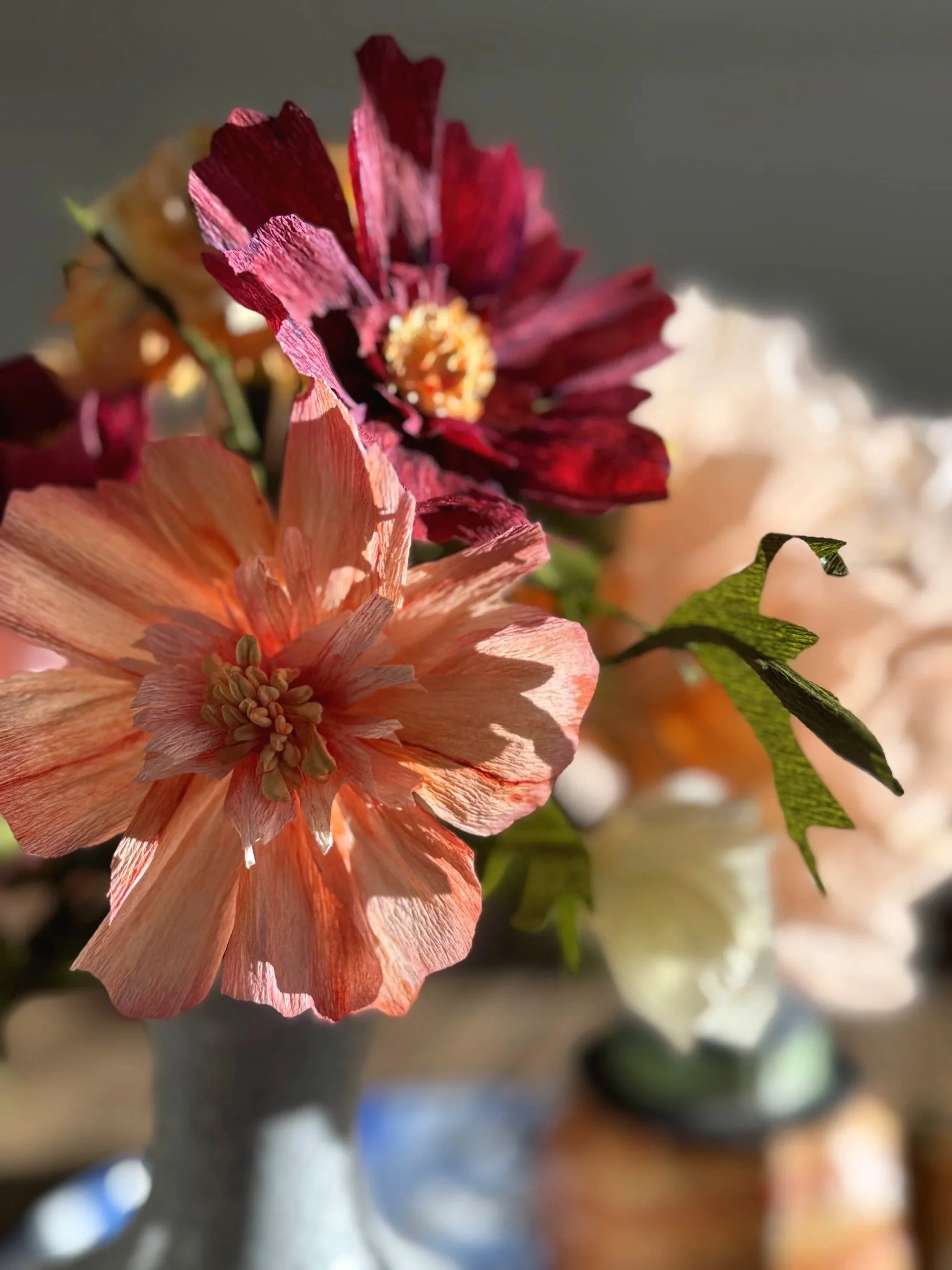 Close-up of artificial flowers in a vase, including a peach-colored flower and a deep red flower, with some green leaves, against a blurred background.