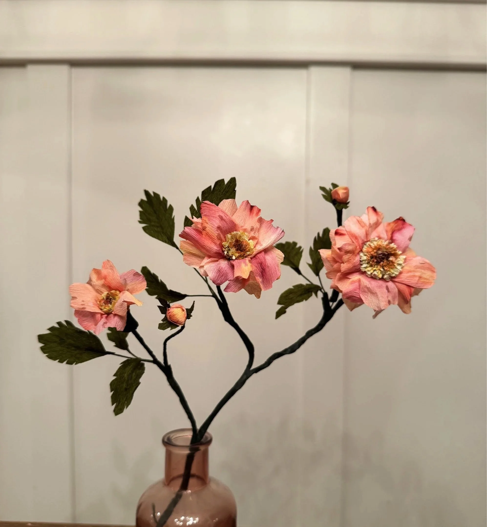 Decorative porcelain vase with pink artificial flowers and green leaves on dark stems, set against a white paneled background.