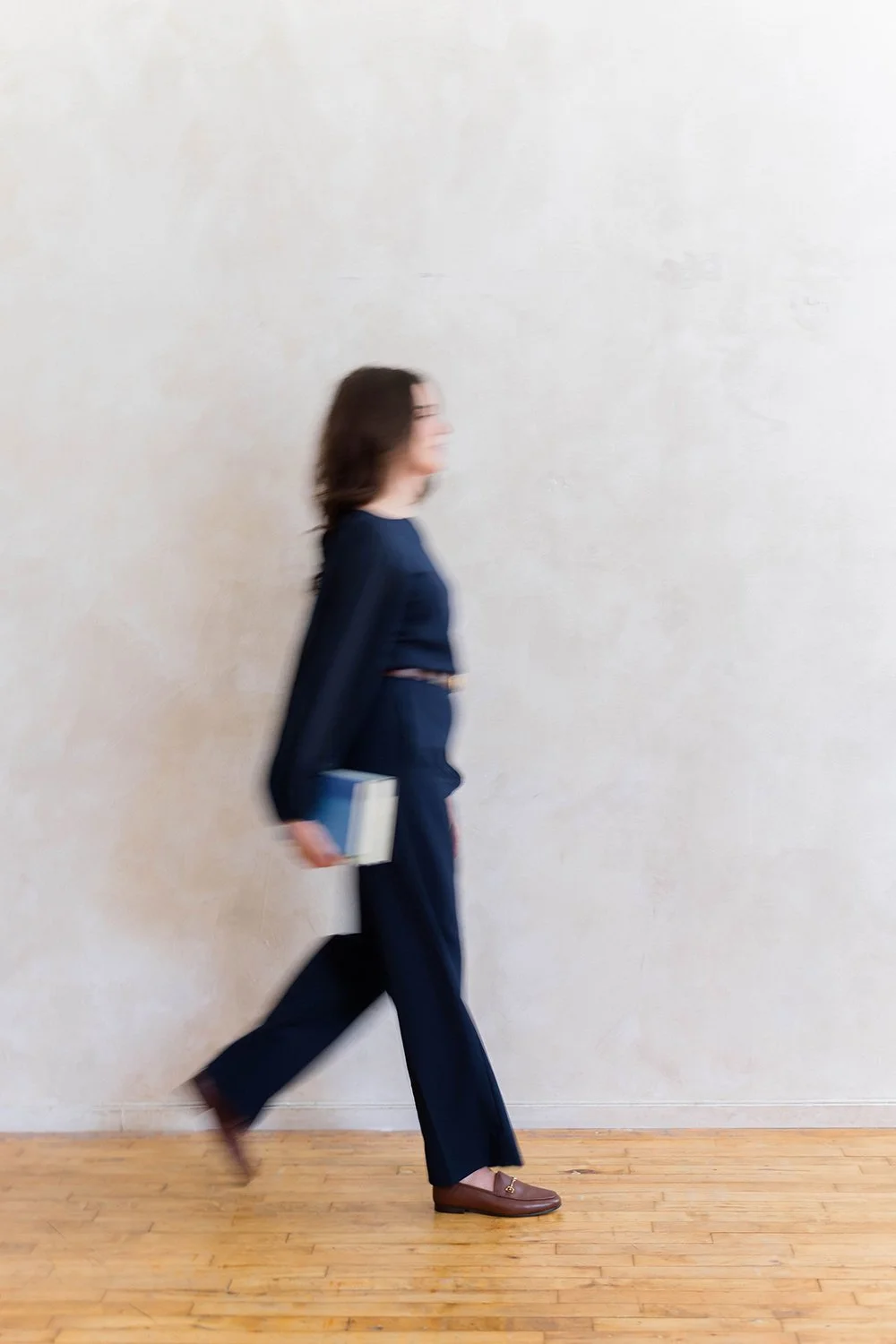 A woman walking indoors with a blurred motion, holding a book in her right hand, against a plain light-colored wall and wooden floor.