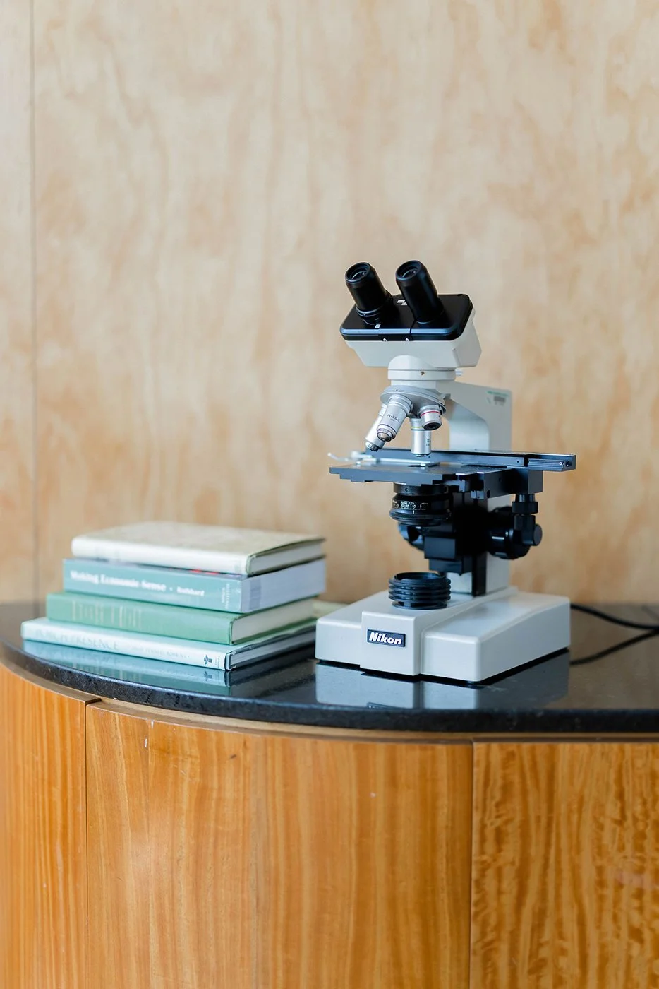 A microscope with black eyepieces and a white body, placed on a wooden table, next to a stack of three books with light green and beige covers, against a wood-paneled wall.