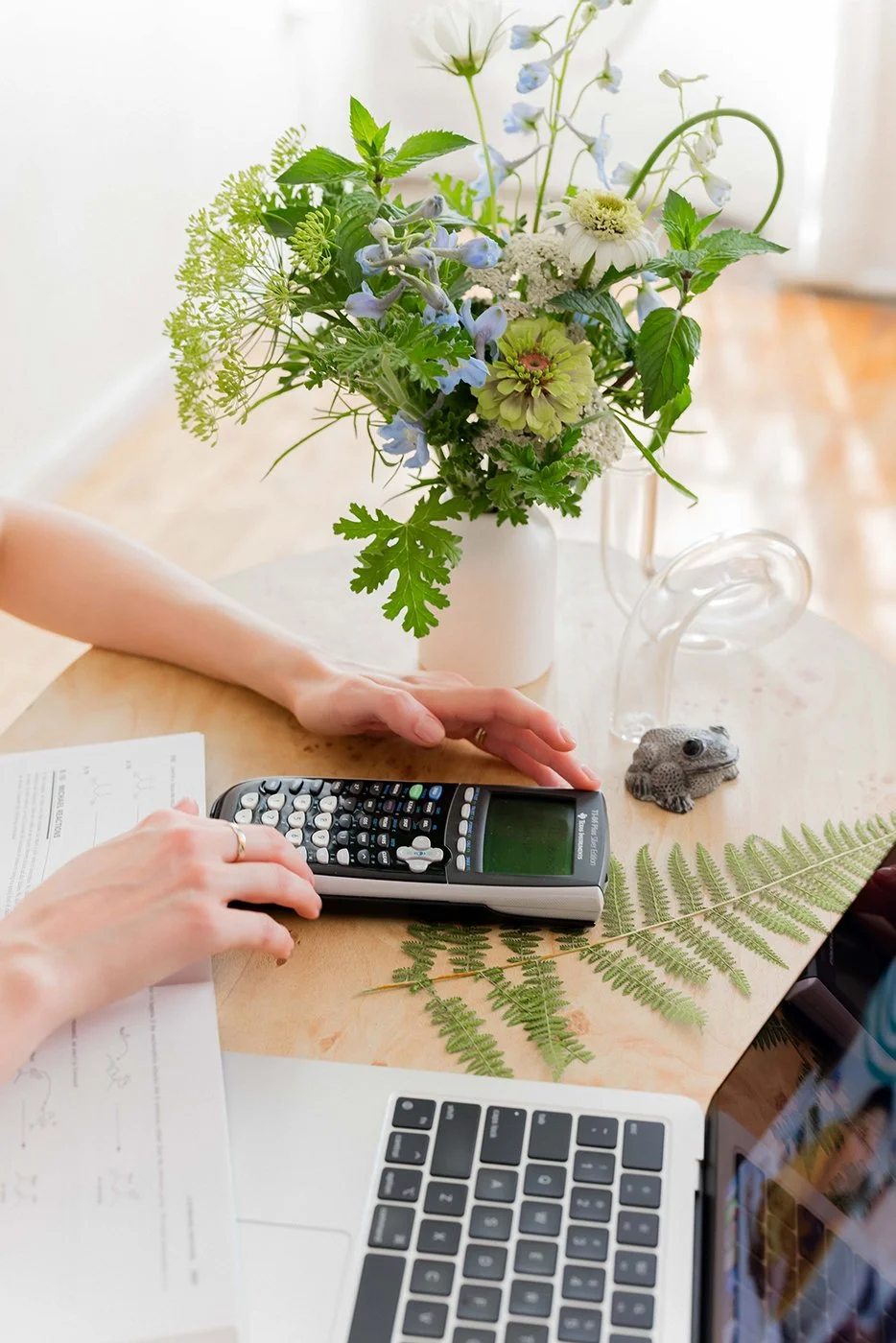 A person's hand operating a scientific calculator on a wooden table with a fern leaf. Items on the table include a notebook, a frog figurine, and a white vase with a bouquet of white and blue flowers and green foliage. Part of a laptop is visible in