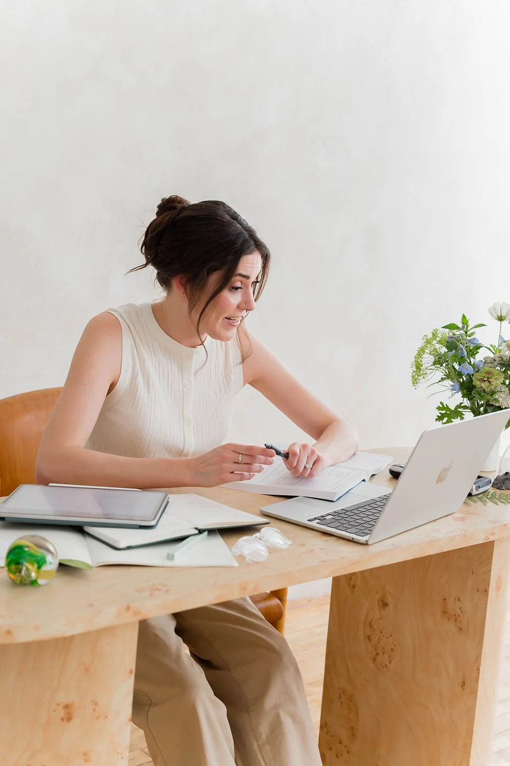 A woman sitting at a wooden desk with open notebooks, a tablet, and a silver laptop, explaining something on a zoom call, with a vase of flowers on the right side.