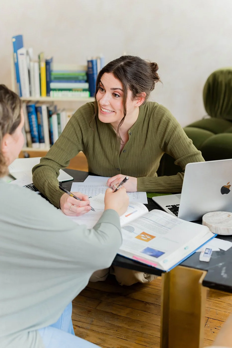 Two women studying chemistry together at a desk with notebooks, papers, laptops, and books, in a study setting.