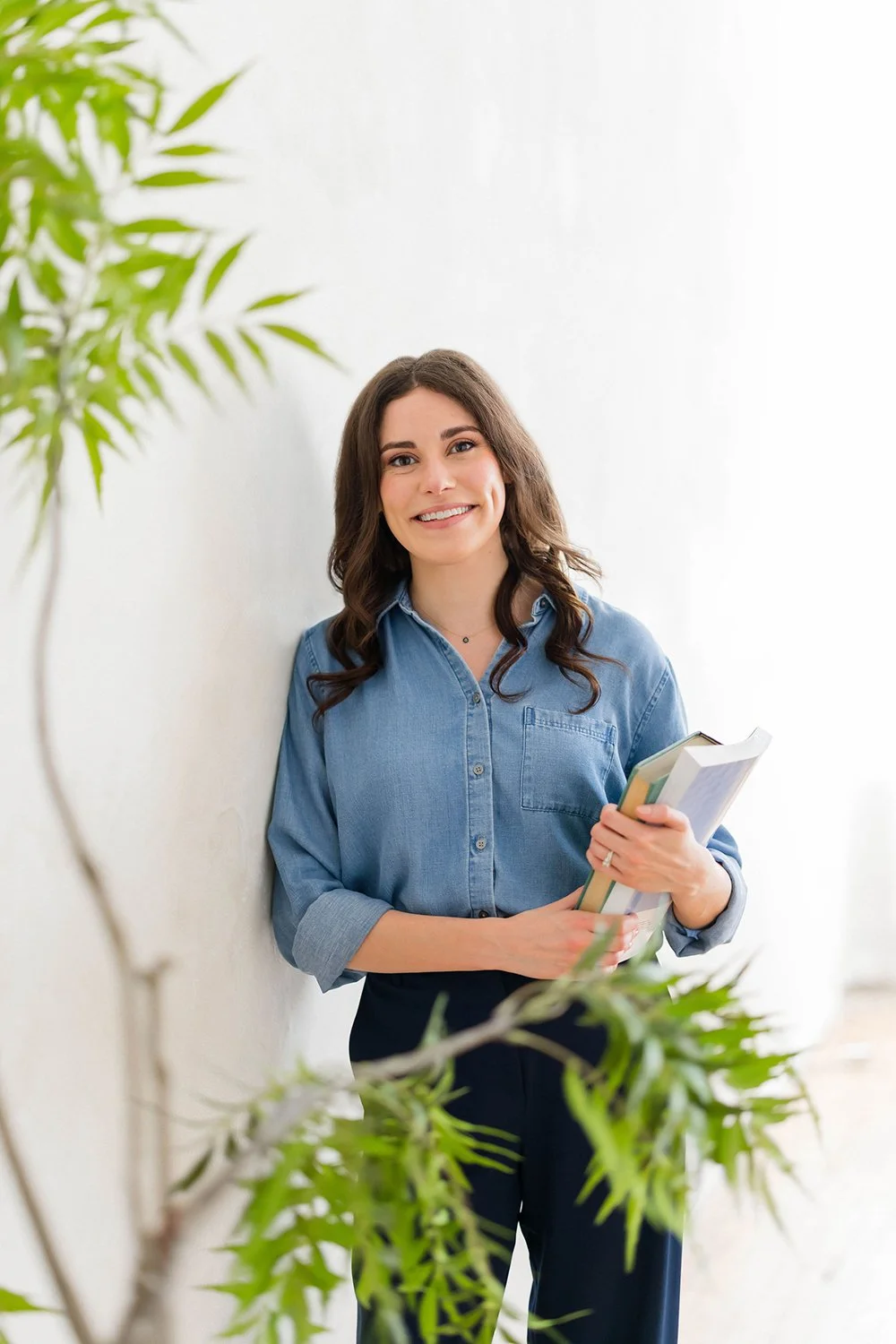 Young woman (Lauren Boulay) with long dark hair, wearing a denim shirt and black pants, smiling while holding books, standing against a white wall with green leaves in the foreground.