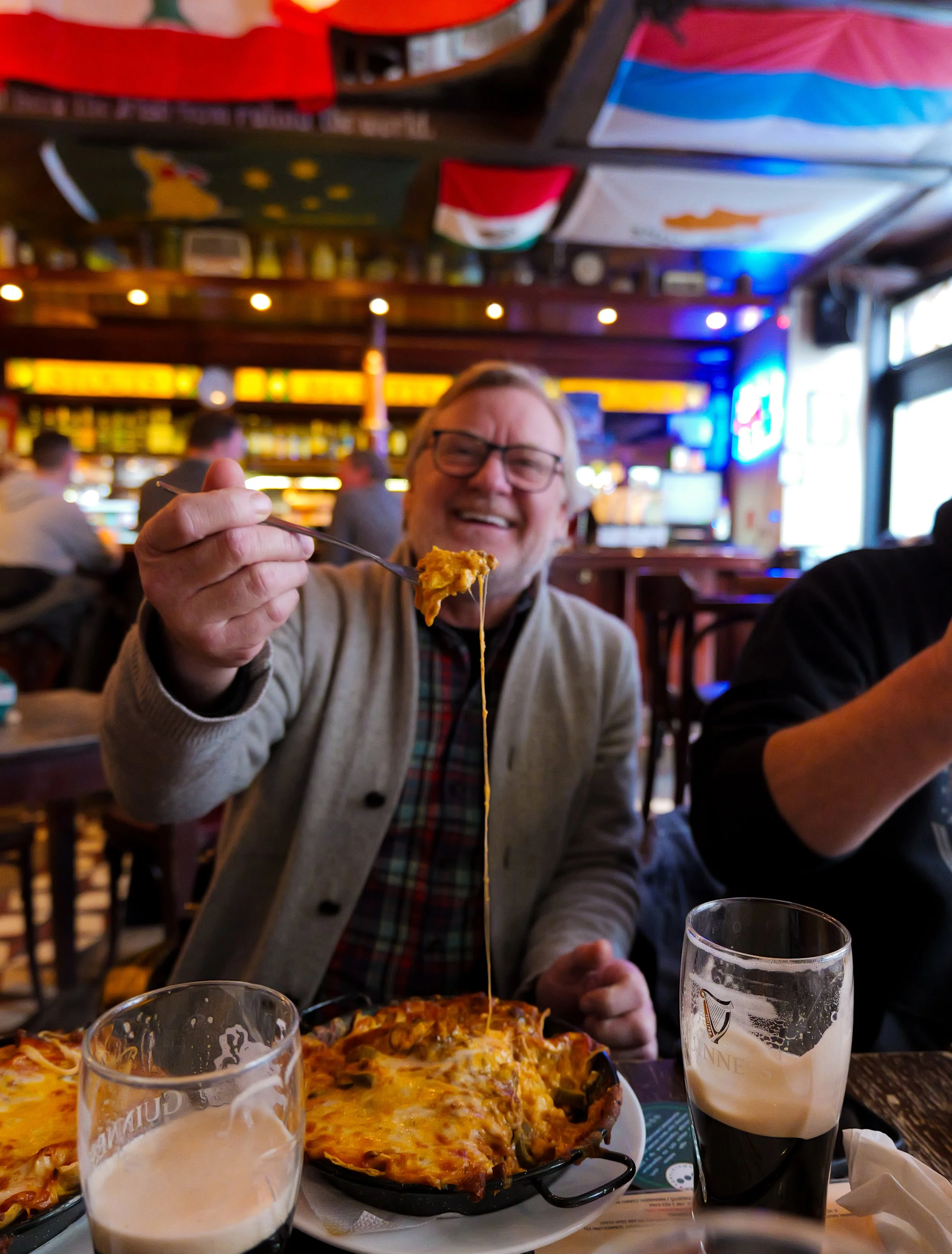 A man with glasses and a beige cardigan smiling while holding a fork with cheesy food, sitting at a table in a lively restaurant. There are pizza and dark beer in front of him.