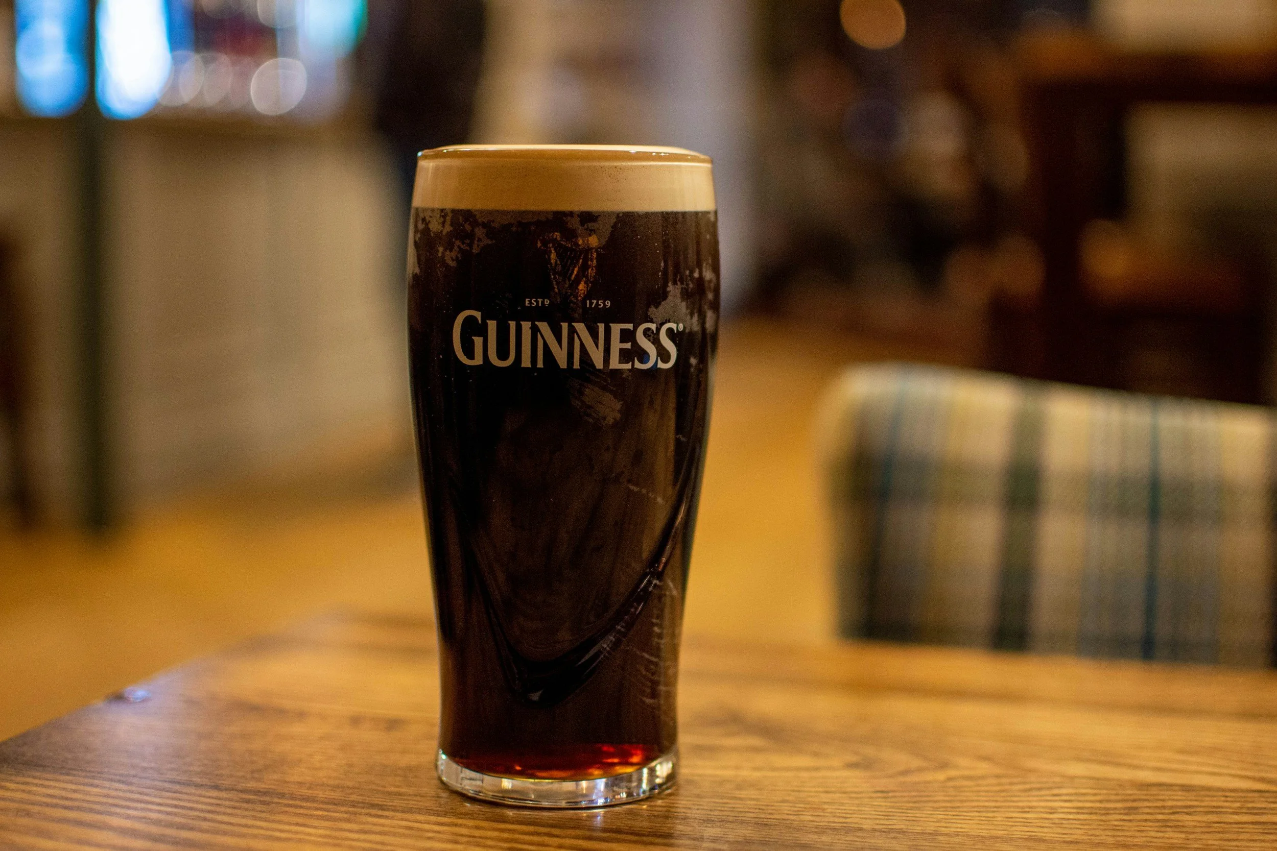 A pint glass of Guinness stout beer on a wooden table in a pub, with a foamy head and dark liquid inside.