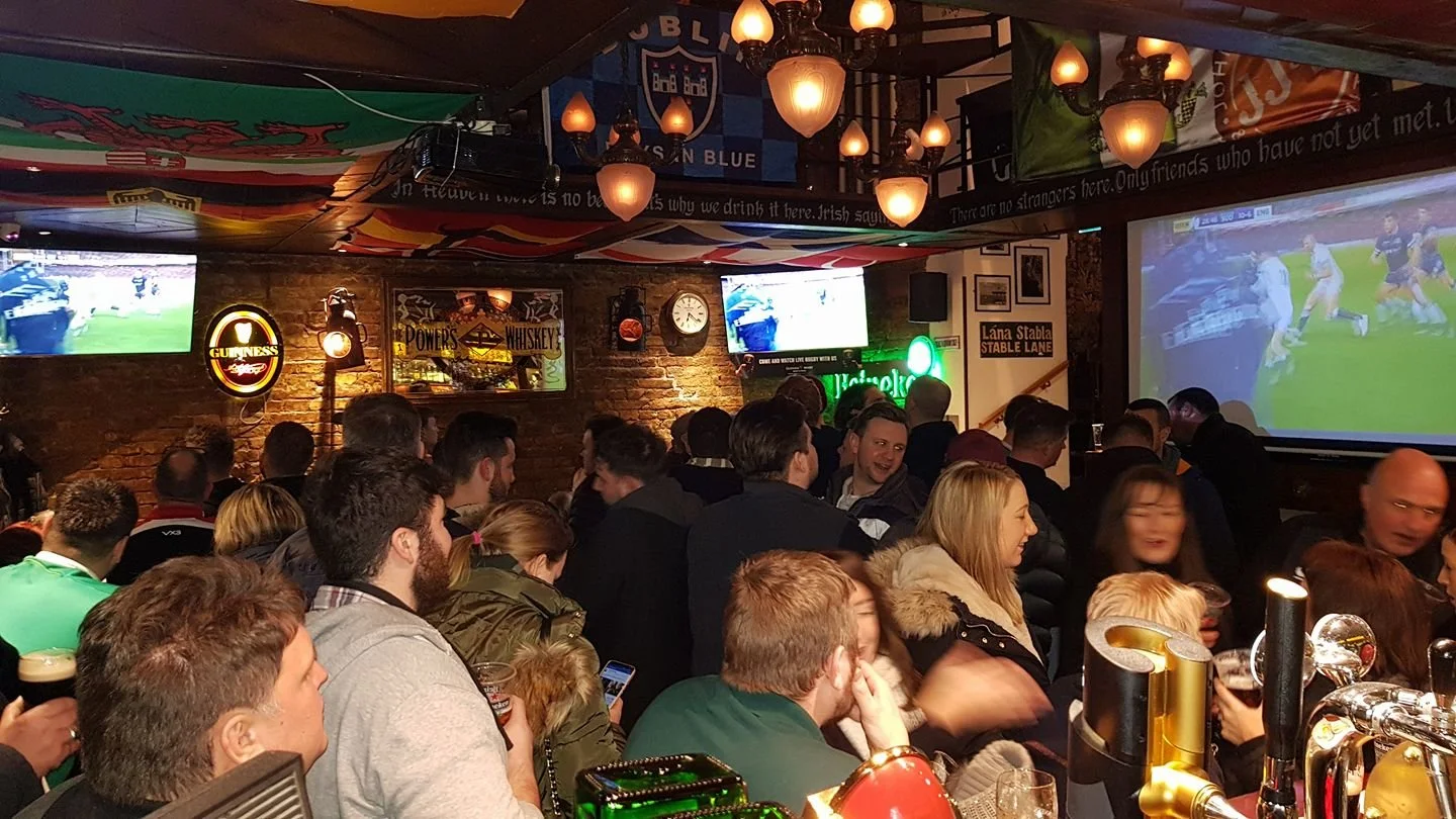 Crowded pub with people watching sports on multiple screens, bar counter with taps, Irish decor, and banners on the ceiling.