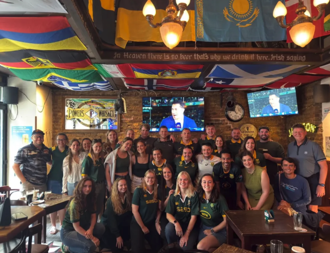 Group of people gathered inside a pub, some wearing sports jerseys, with television screens showing a man speaking, flags hanging from the ceiling, and a sign referencing whiskey.