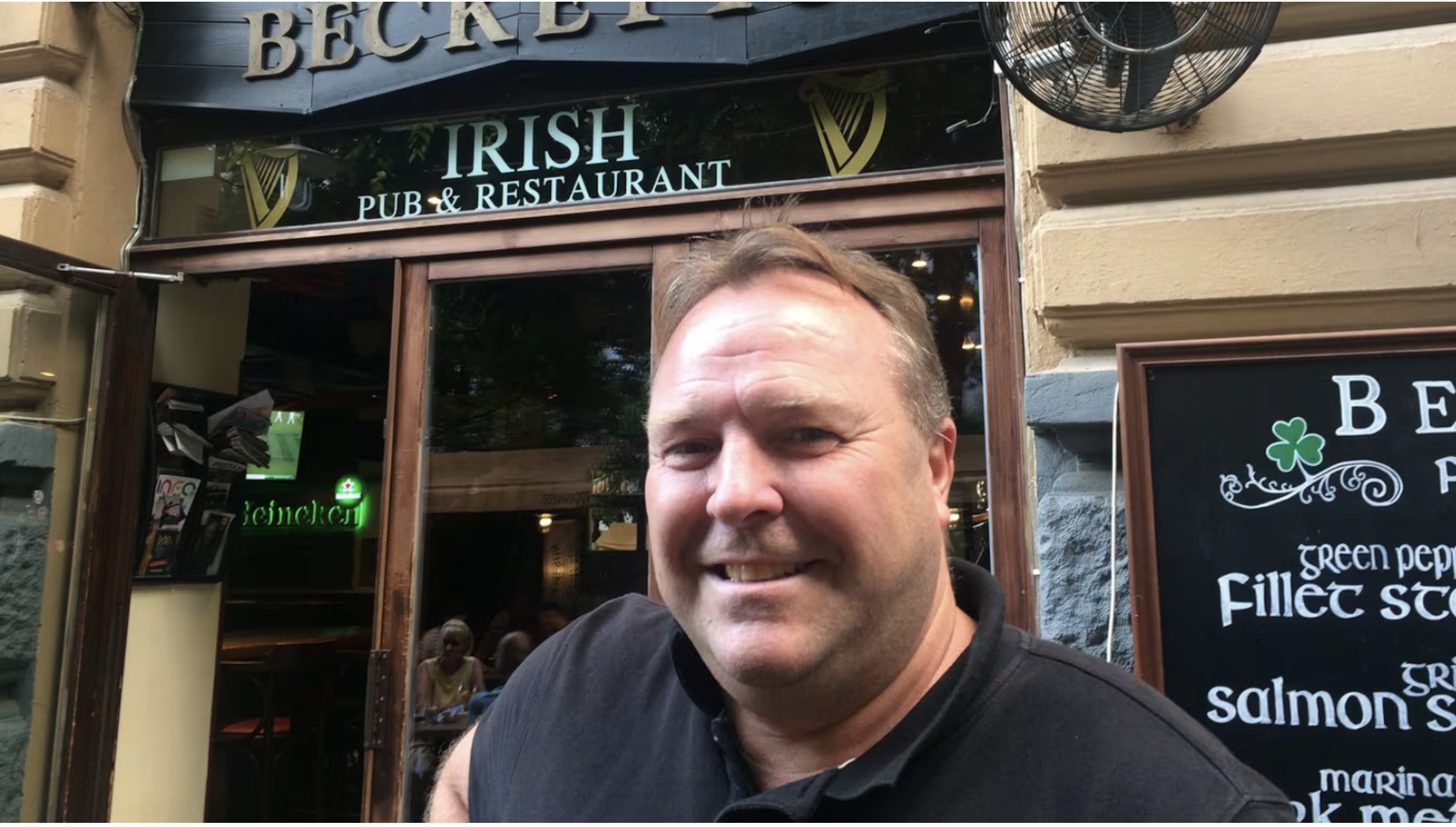 Man smiling standing outside an Irish pub and restaurant with a sign for Guinness and a chalkboard menu listing various dishes.