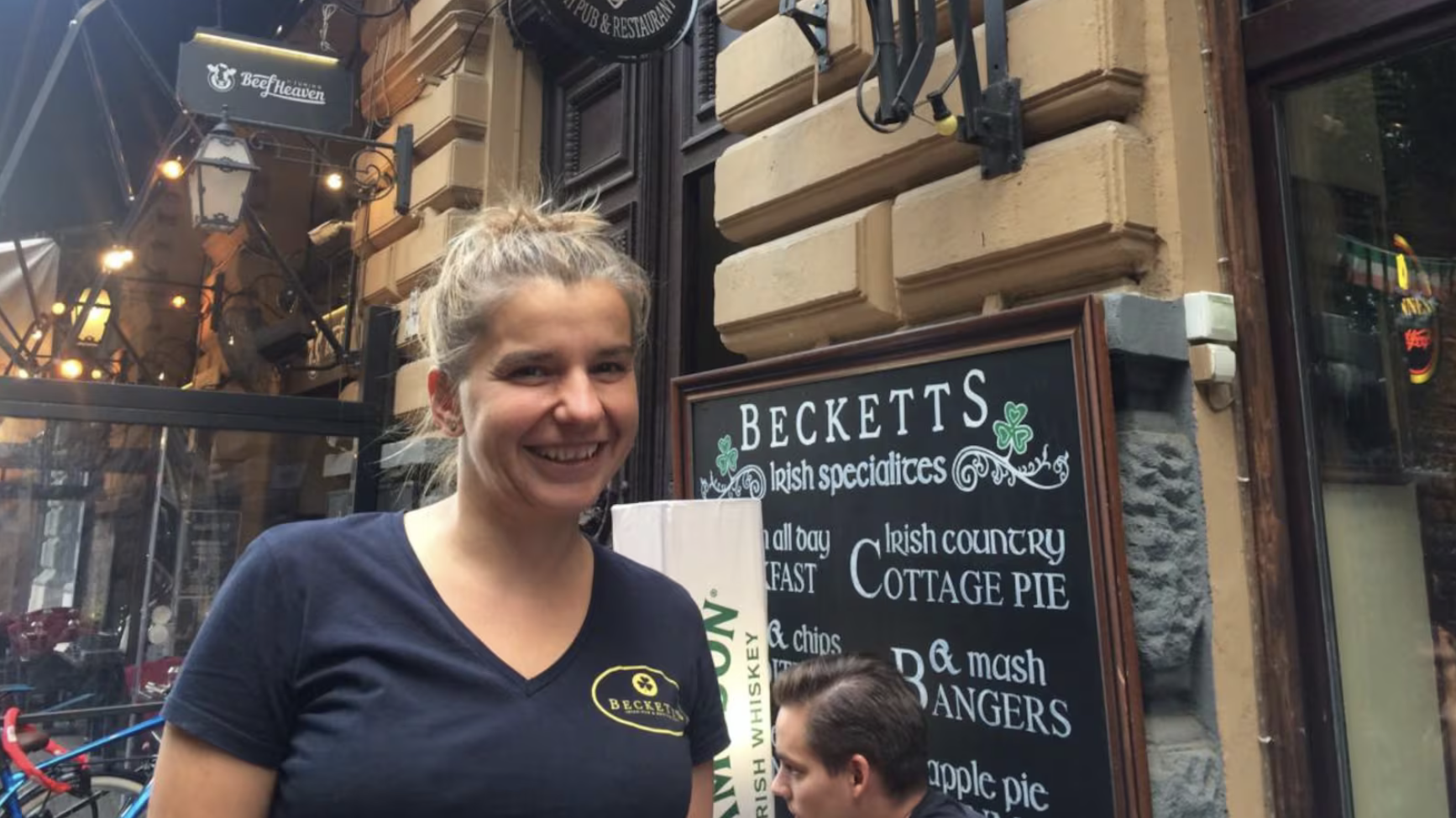 A smiling woman stands outside a restaurant with signs advertising Irish specialties and a menu. She is wearing a black T-shirt with the word 'Beckett's' on it.