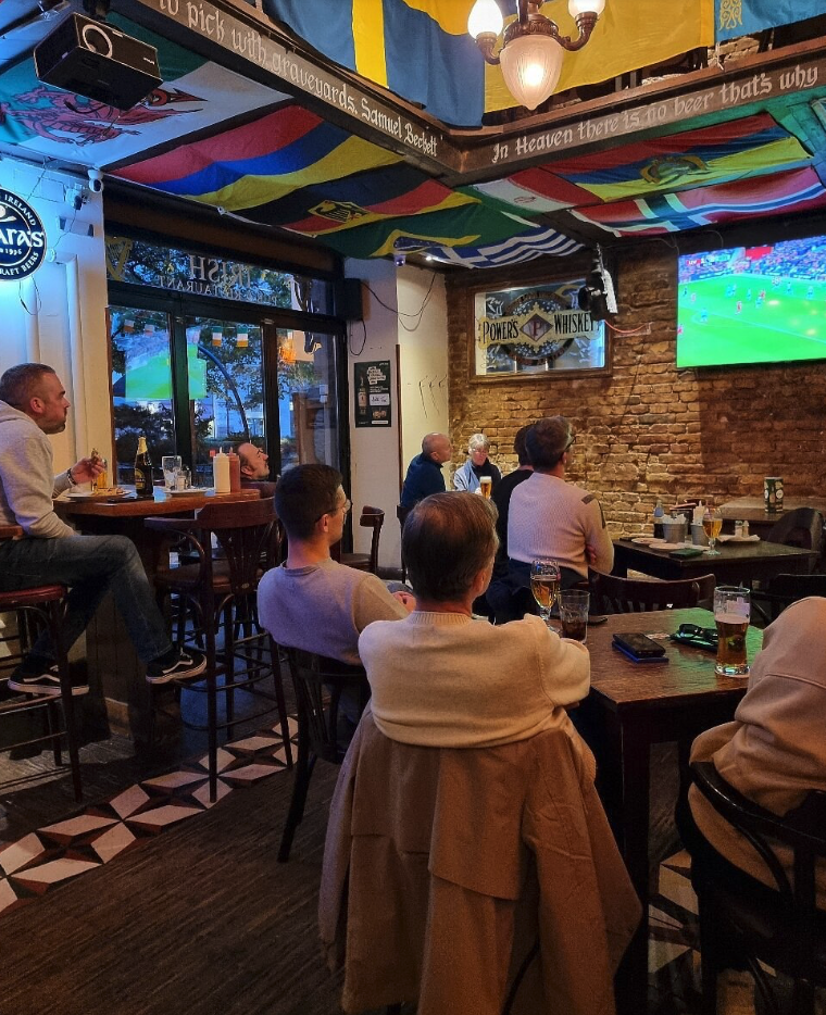 Group of people watching a soccer game on television in a pub, with a brick wall, colorful flags on the ceiling, and wooden tables.