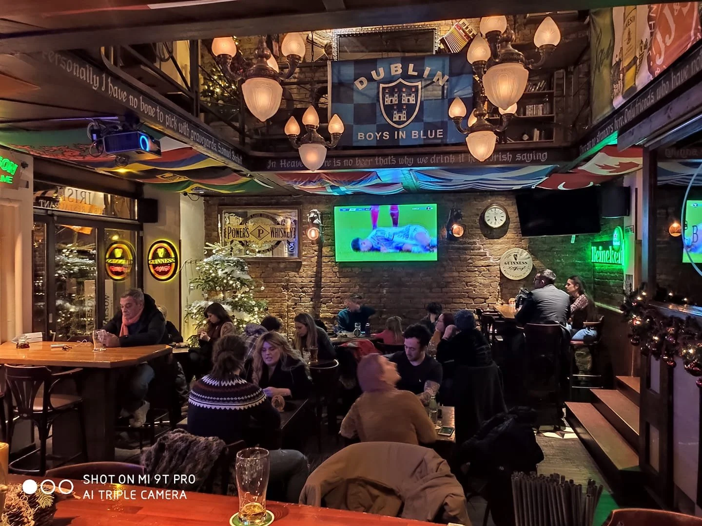Interior of a lively pub decorated for Christmas with a Christmas tree, banners, and ornaments. Patrons are watching a sports game on multiple TVs, some drinking and socializing. Signs for Guinness and Heineken are visible, and a large Dublin Boys in Blue banner hangs from the ceiling. The atmosphere is cozy with warm lighting and brick walls.