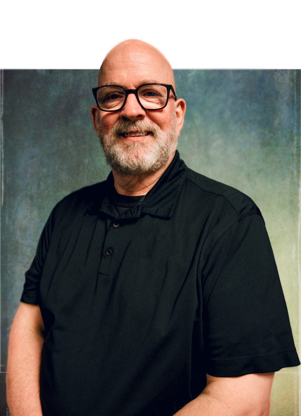 A middle-aged man with glasses and a beard smiling, dressed in a black polo shirt, standing against a textured green background.