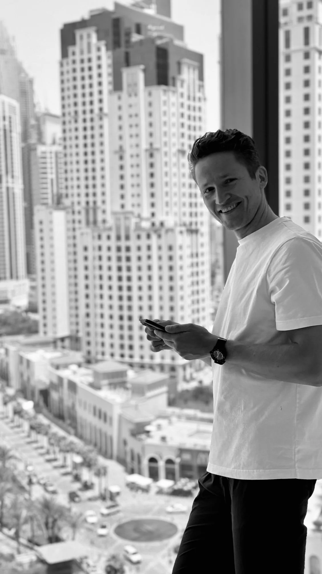 A man smiling and looking at the camera while holding a smartphone, standing in front of a large window with city high-rise buildings in the background.