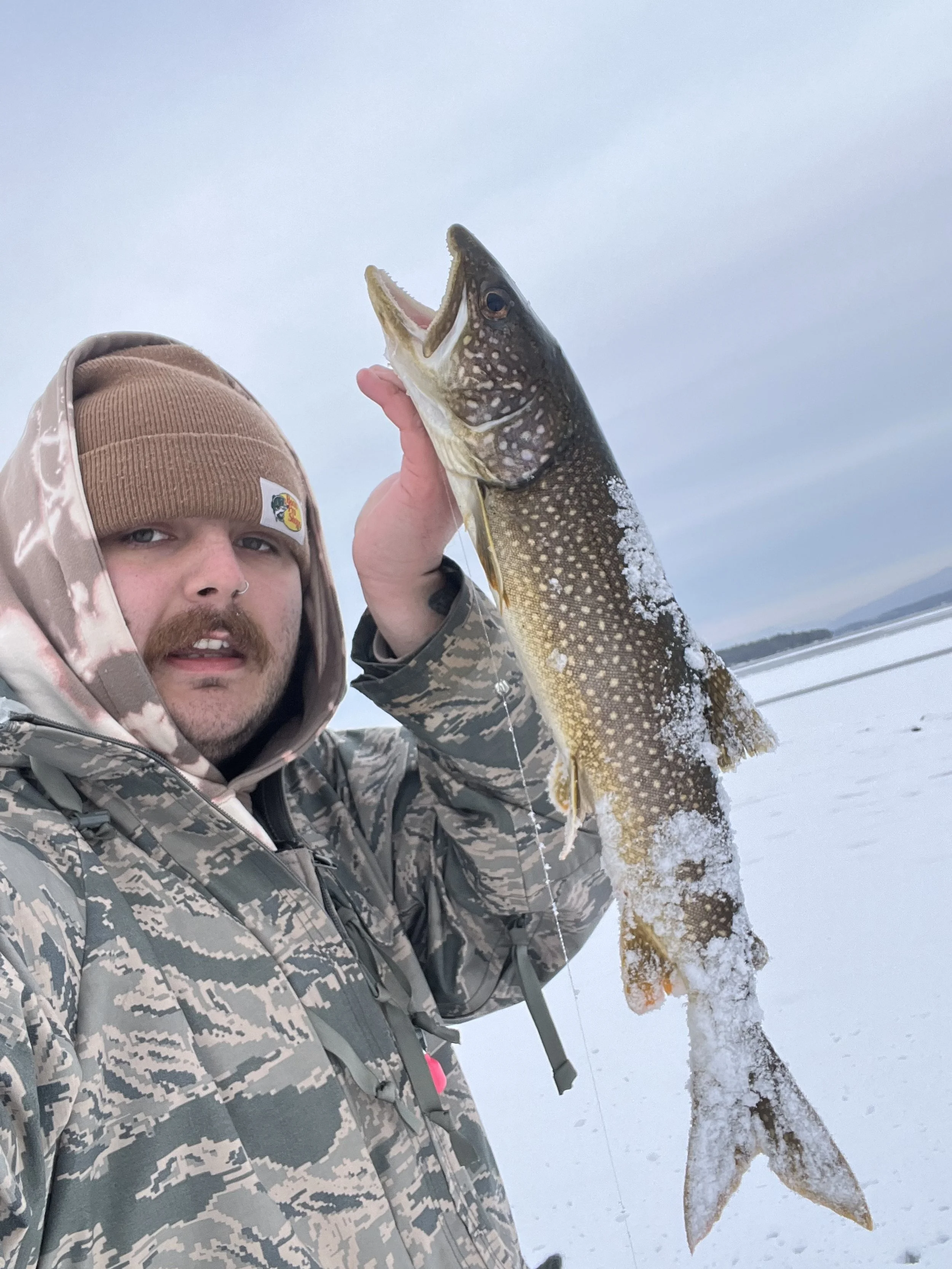 A man in camouflage clothing holding a large fish with a snowy landscape and lake in the background.