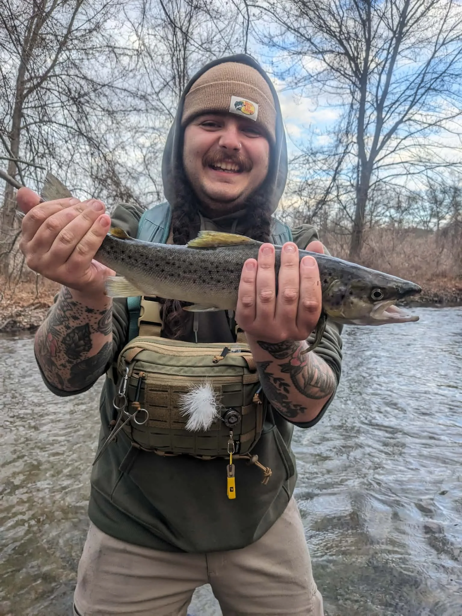 A smiling man with tattoos on his arms holding a large fish outdoors near a river, wearing a beanie and jacket, with a background of trees and a partly cloudy sky.