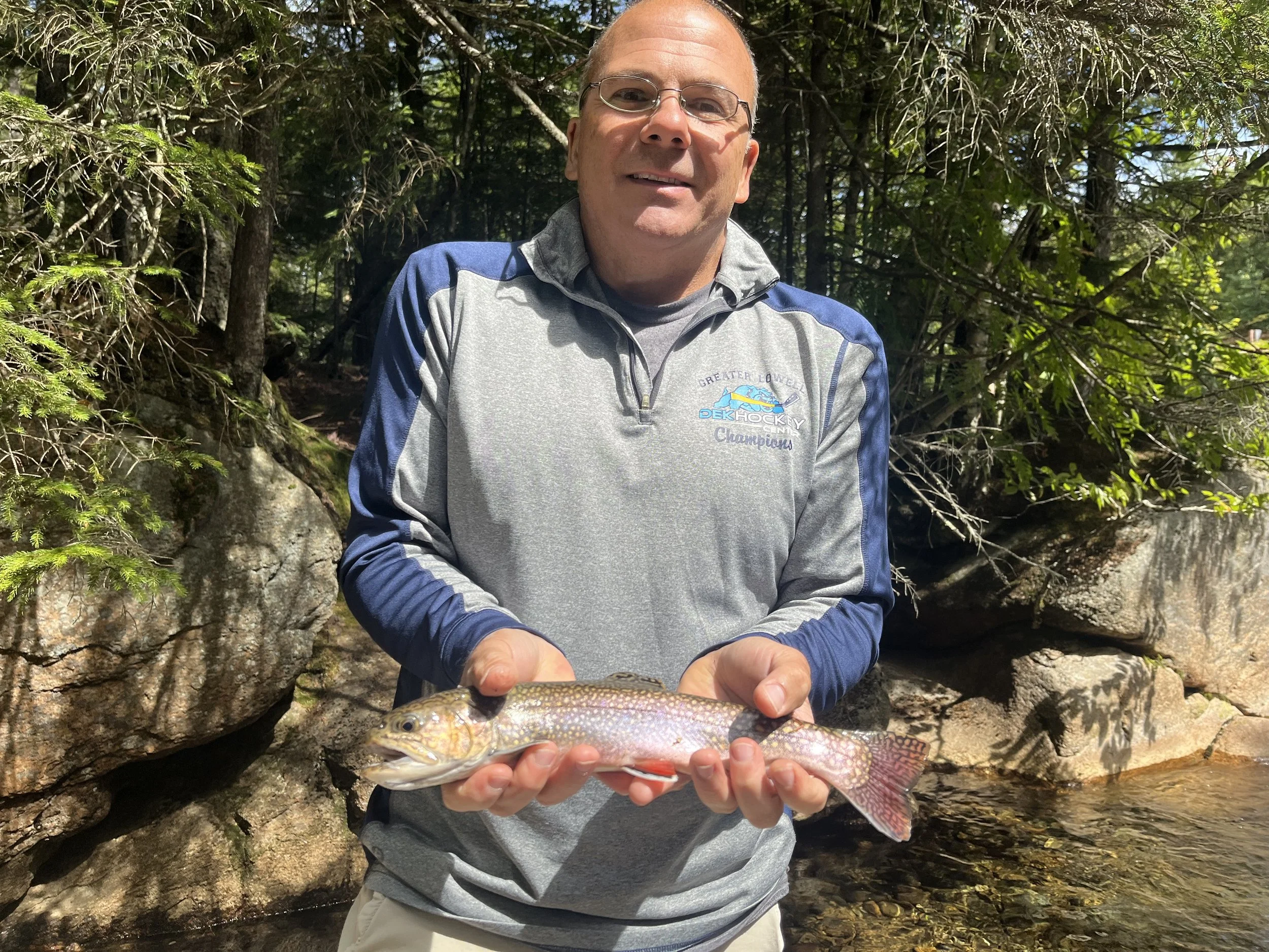A man holding a brook trout outdoors near rocks and trees, smiling at the camera.