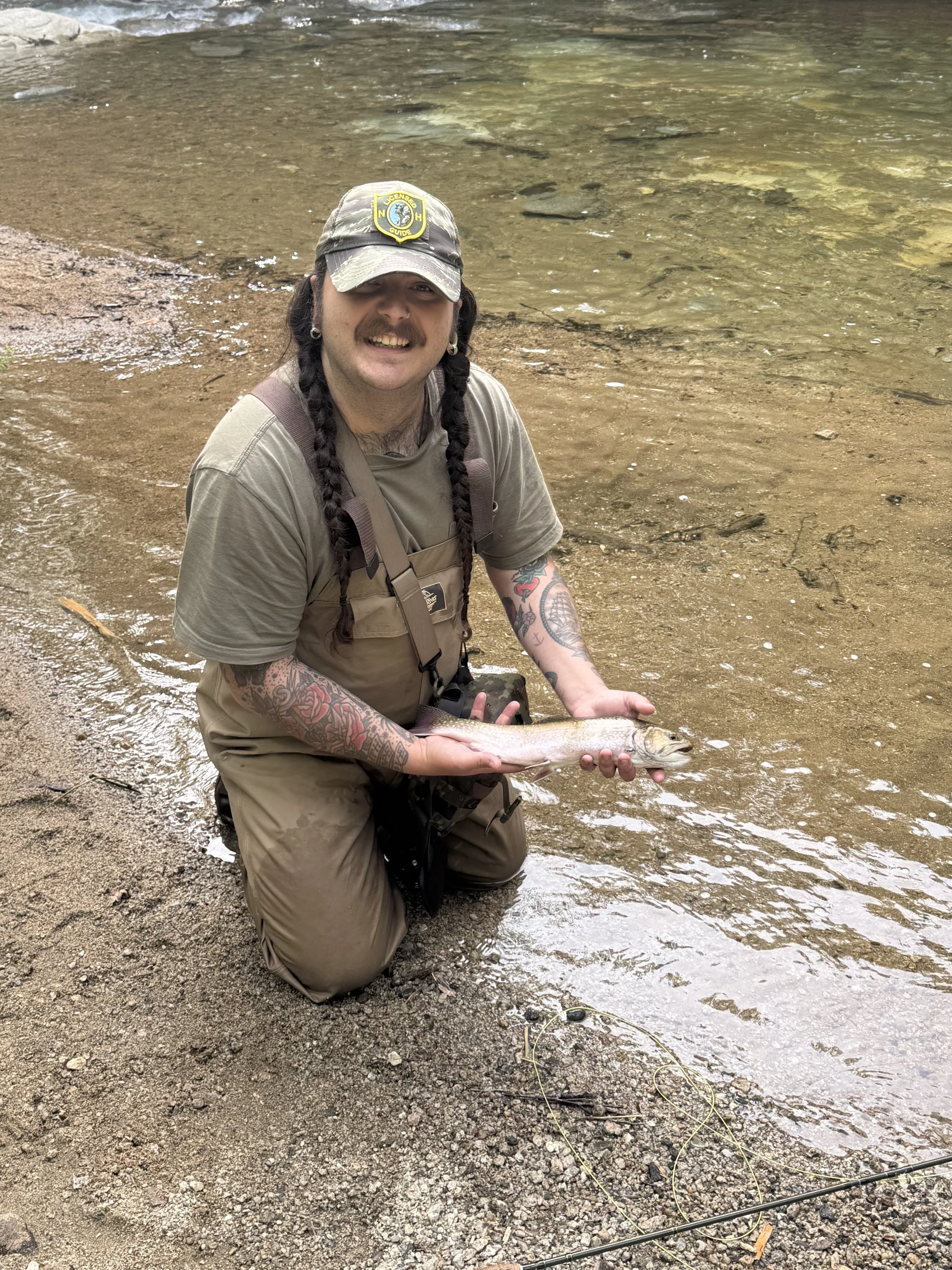 A man kneeling in shallow water at a riverbank, holding a fish, and smiling at the camera. He has long braided hair, tattoos, and is wearing a camouflage cap, khaki clothes, and a fishing vest.