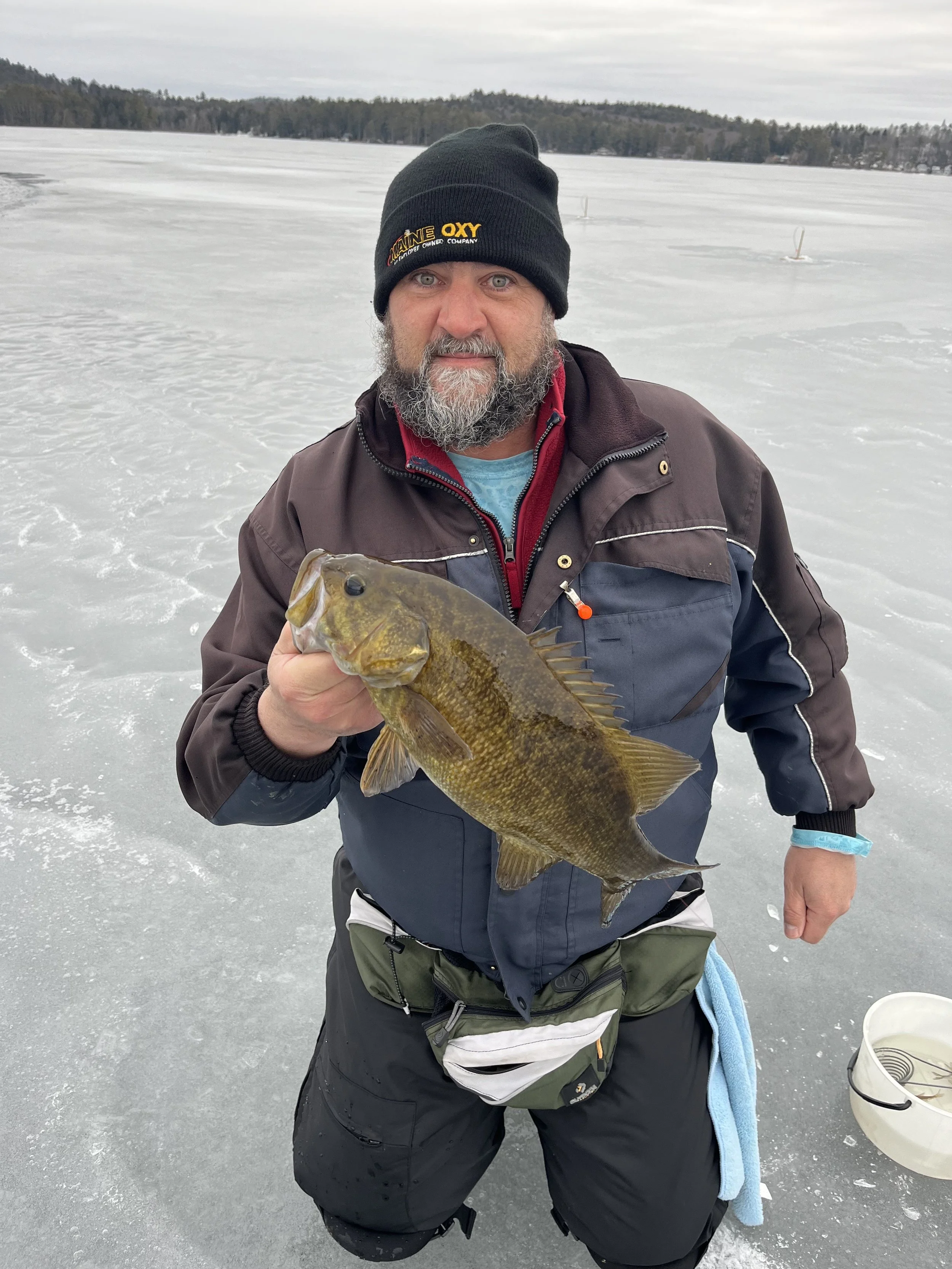 Man in winter clothing and black beanie holding a fish on a frozen lake with trees in the background.