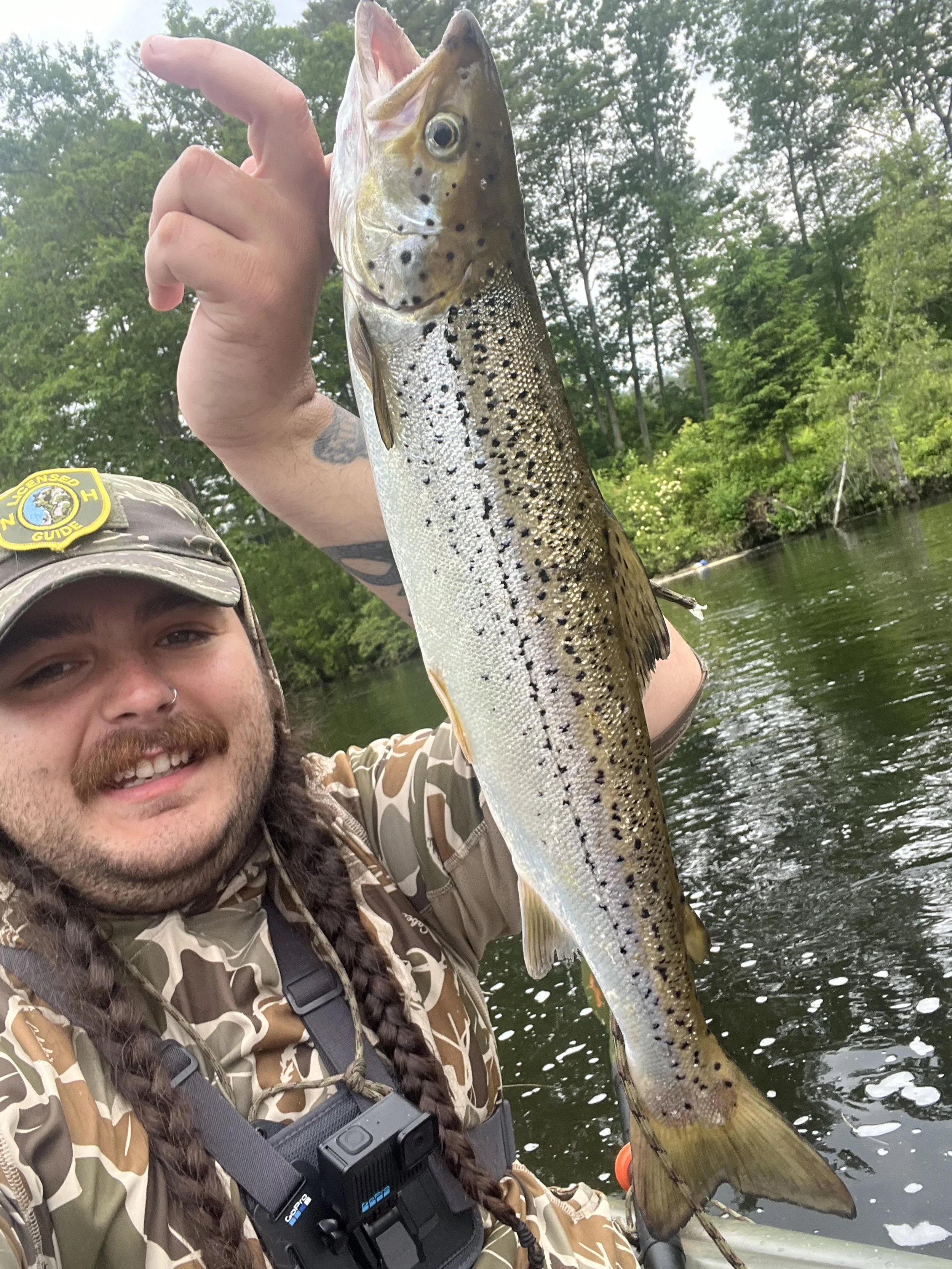 A man holding a large fish, likely a salmon, on a fishing trip in a lush green outdoor setting.