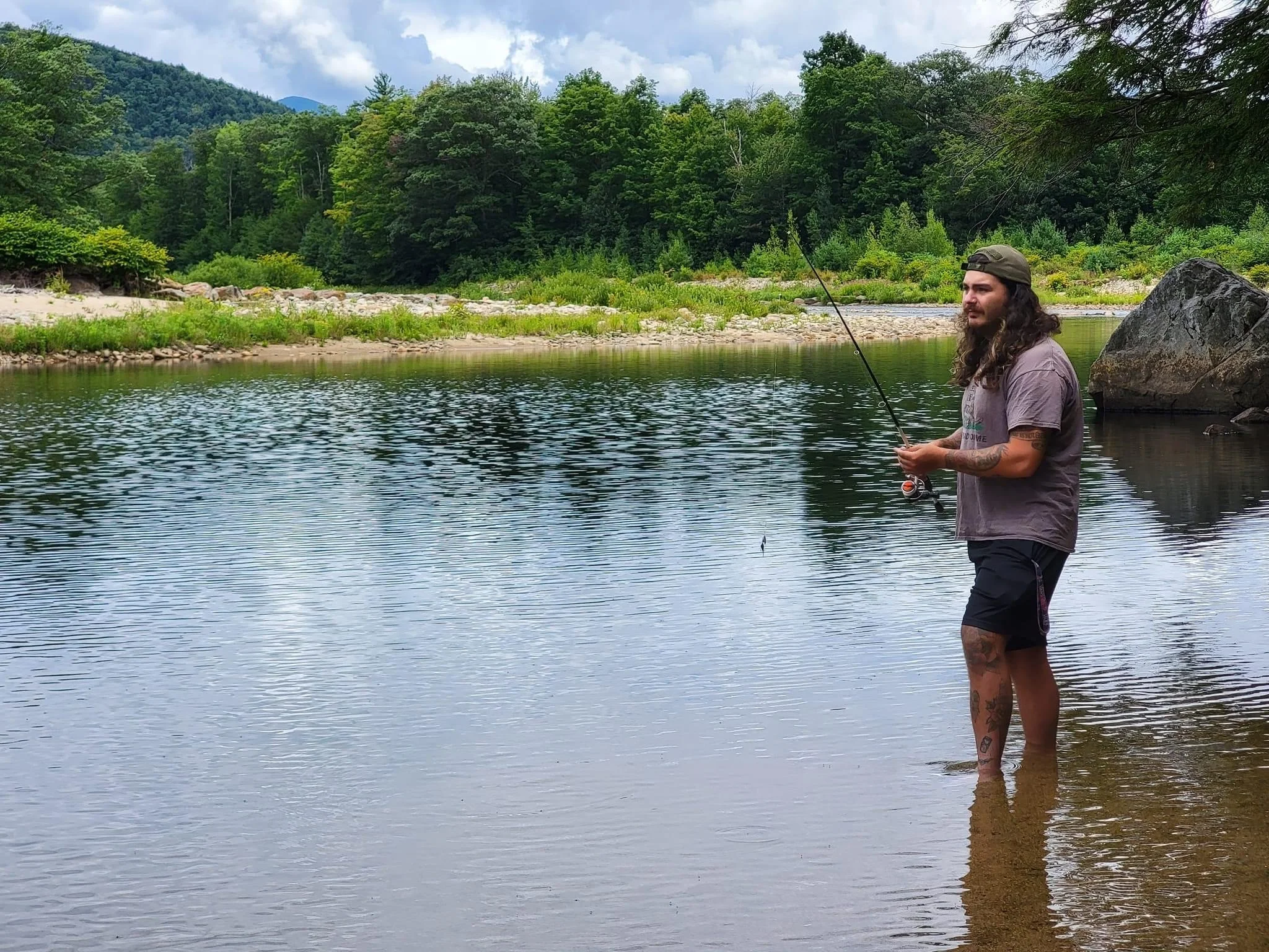 Man fishing in a river with green trees and mountains in the background.