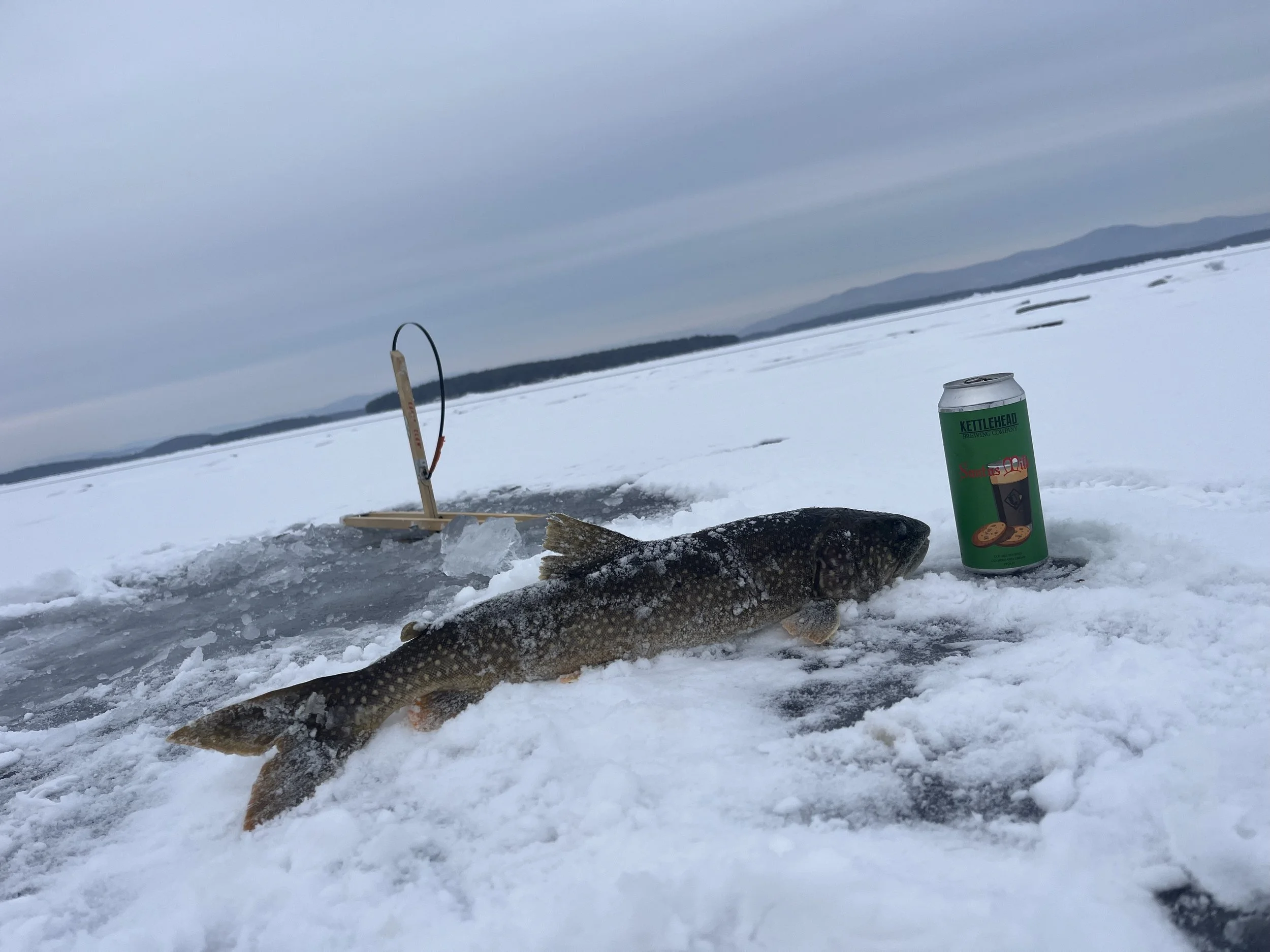 A fish caught on ice with a fish tickler tool, a can of Kettlehead beer, and a mountain landscape in the background.