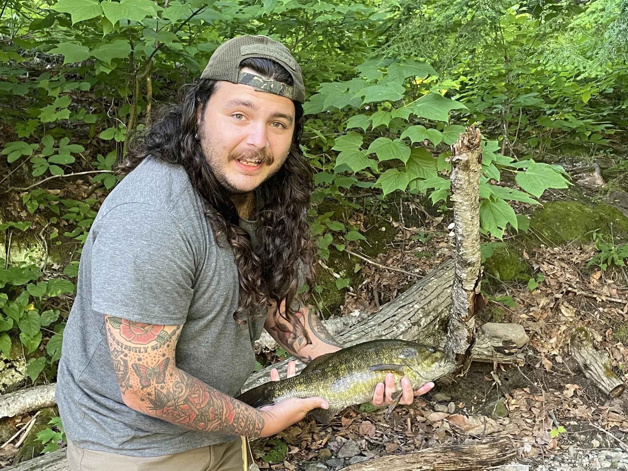 A man with long curly hair, wearing a gray t-shirt and a backwards baseball cap, holding a fish in a wooded area surrounded by green leaves and a fallen log.