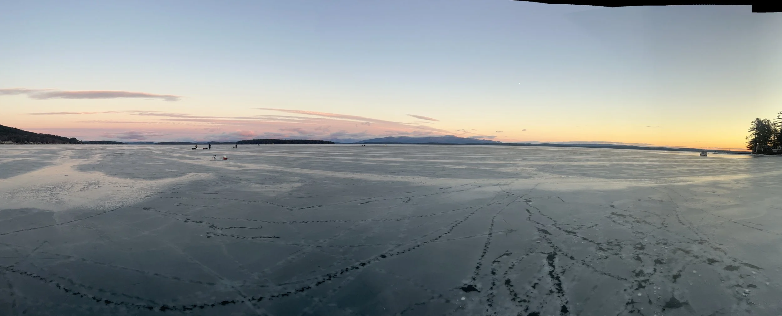 Frozen lake with ice fishing holes and footprints, trees and mountains in the distance at sunset.