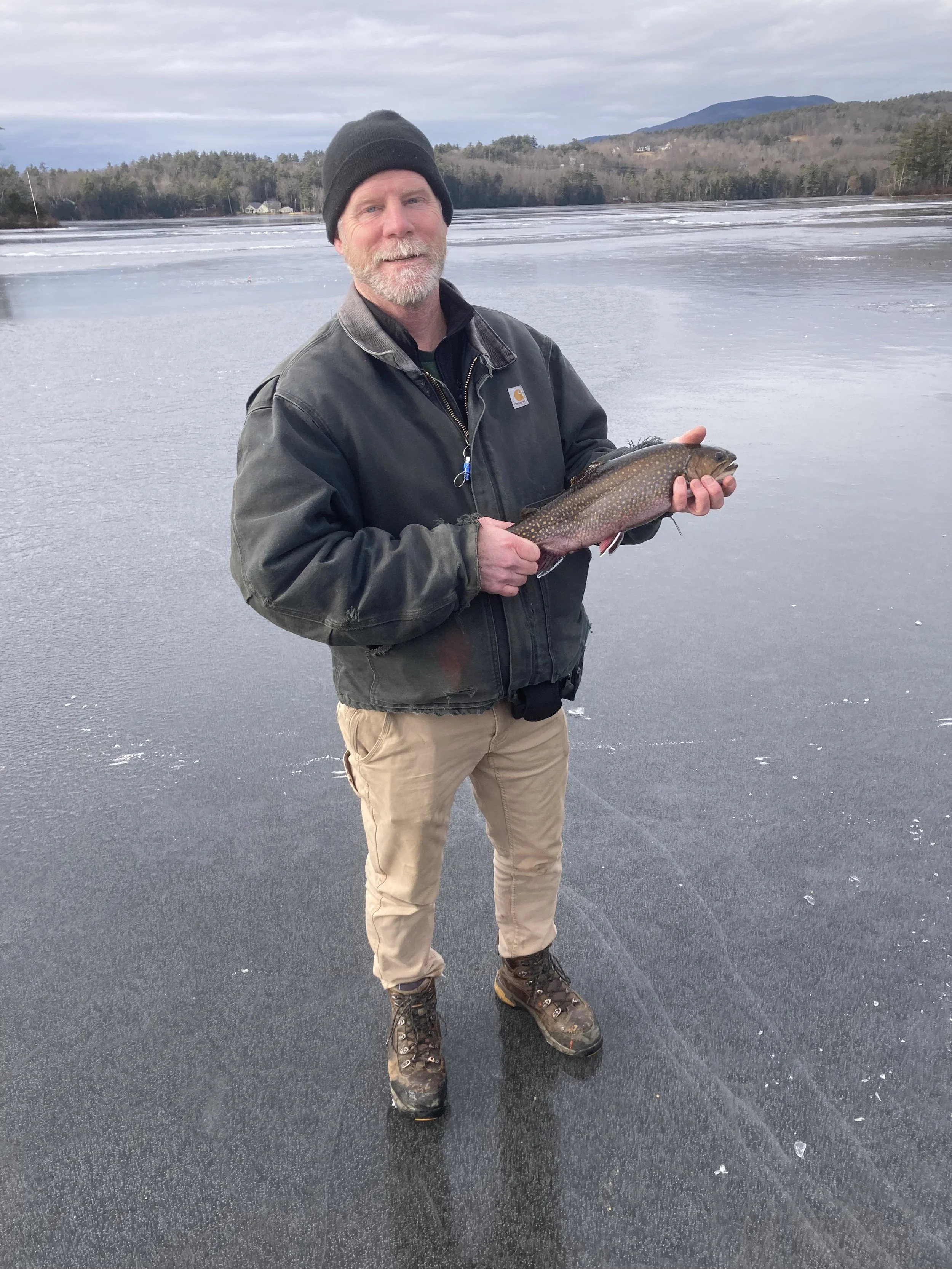 Man standing on a frozen lake holding a fish, wearing a black beanie, black jacket, beige pants, and hiking boots, with a background of trees, hills, and a cloudy sky.
