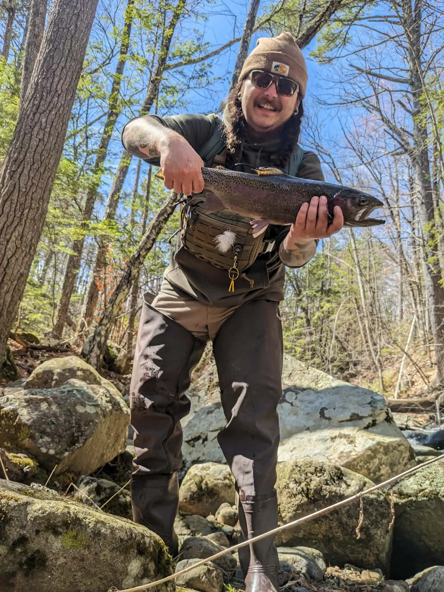 A man with sunglasses, a brown beanie, long hair, and a beard holding a large rainbow trout fish in a forested area with rocks and trees under a clear blue sky.