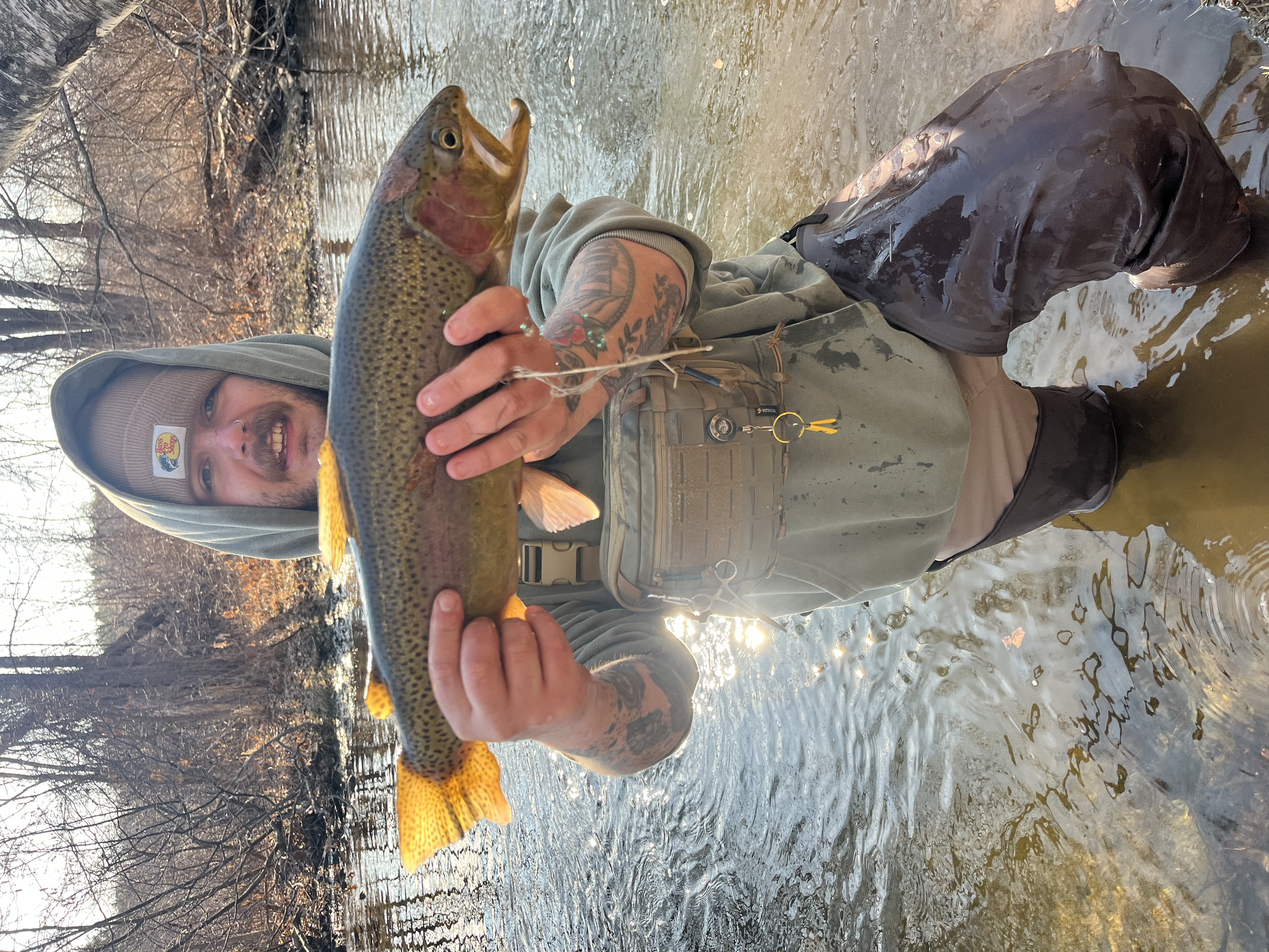A man wearing a hoodie and chest waders standing in a river, holding a large fish, likely a trout, with a smile on his face.