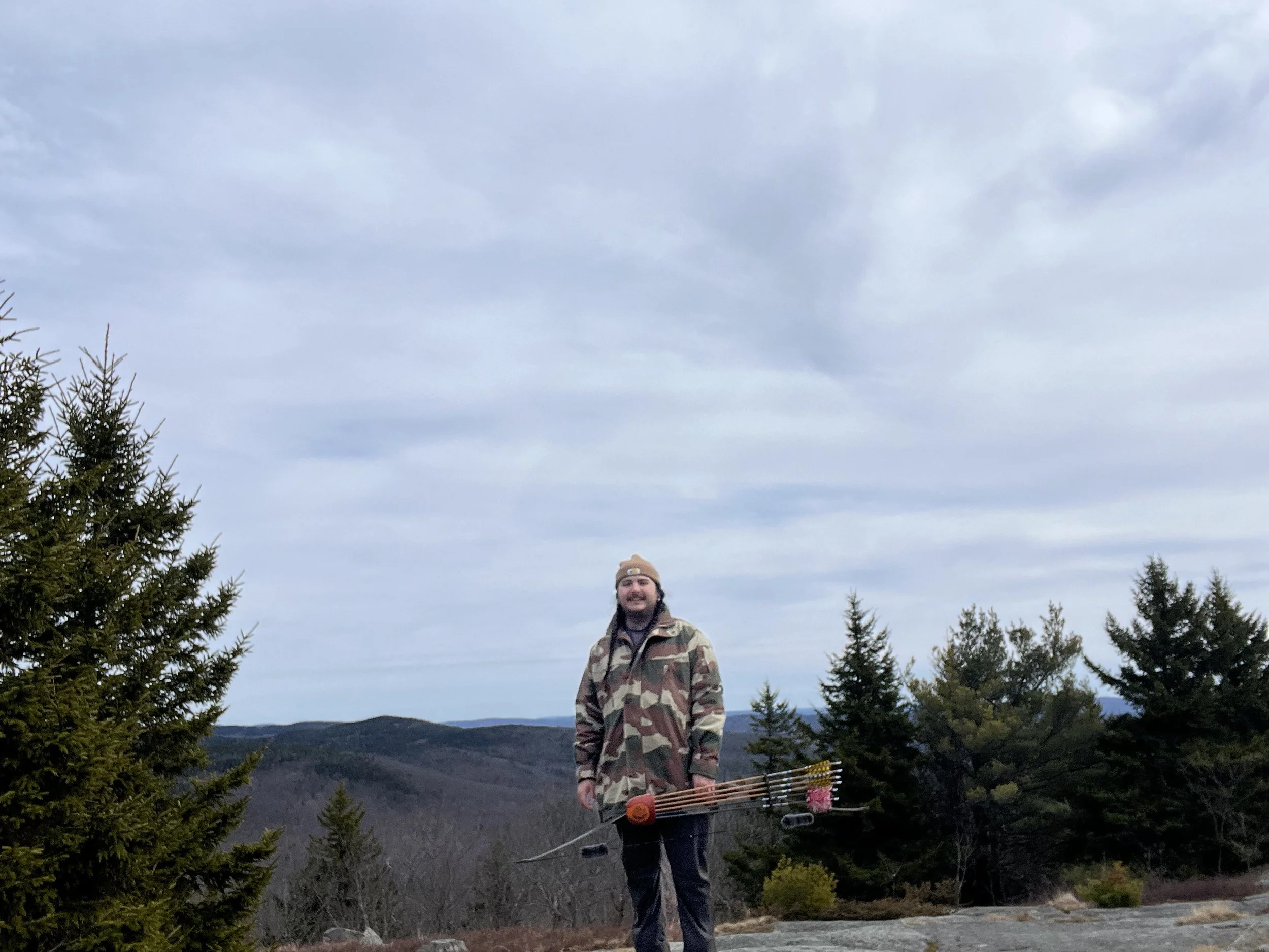 A man standing outdoors on rocky terrain, smiling, dressed in camouflage jacket, holding a bow with arrows attached, with evergreen trees and mountain hills in the background under a cloudy sky.