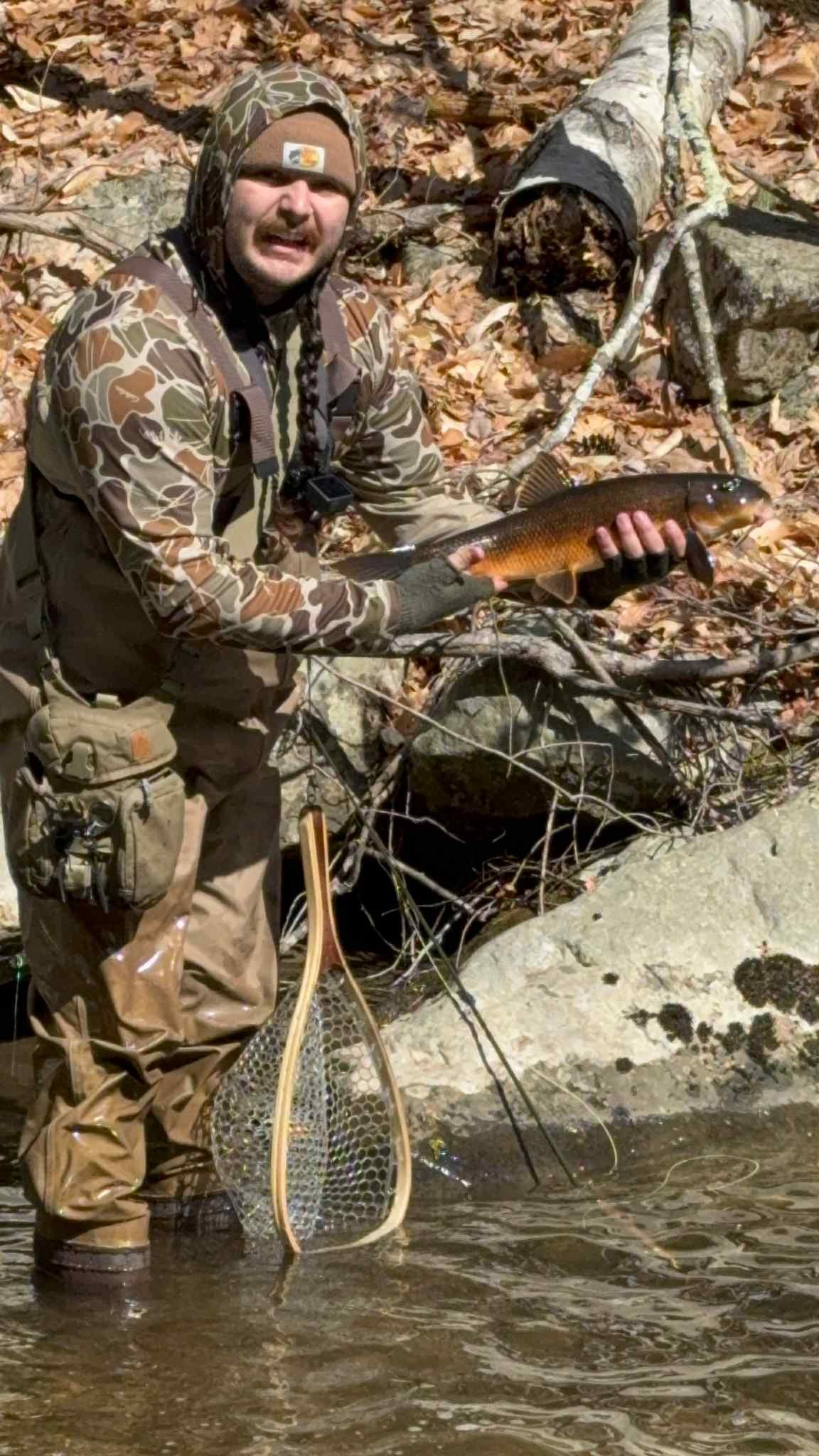 Man in camouflage clothing holding a fish beside a riverbank with fallen leaves and rocks.