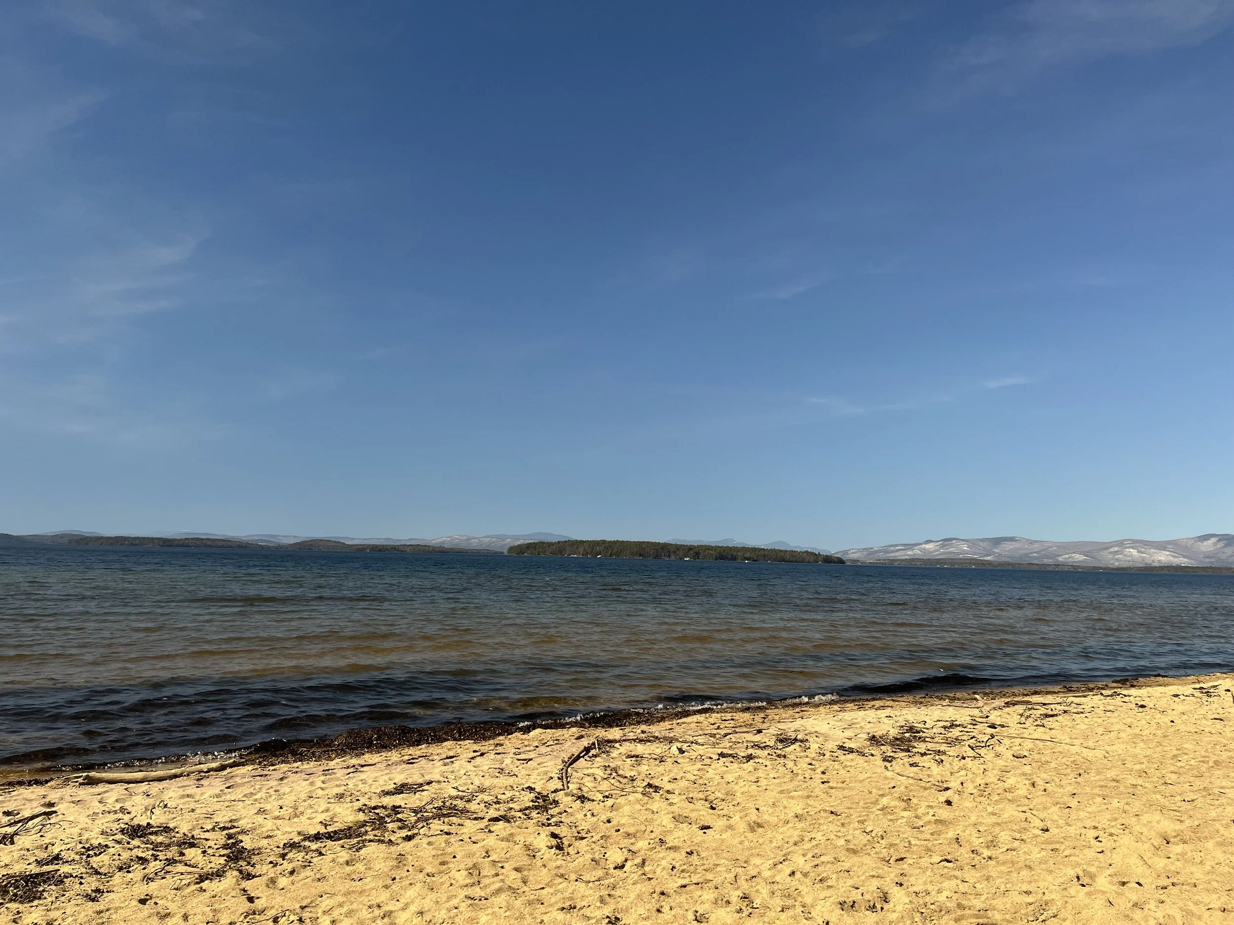A sandy beach with gentle waves in a lake or sea, distant islands, and snow-capped mountains under a clear blue sky.