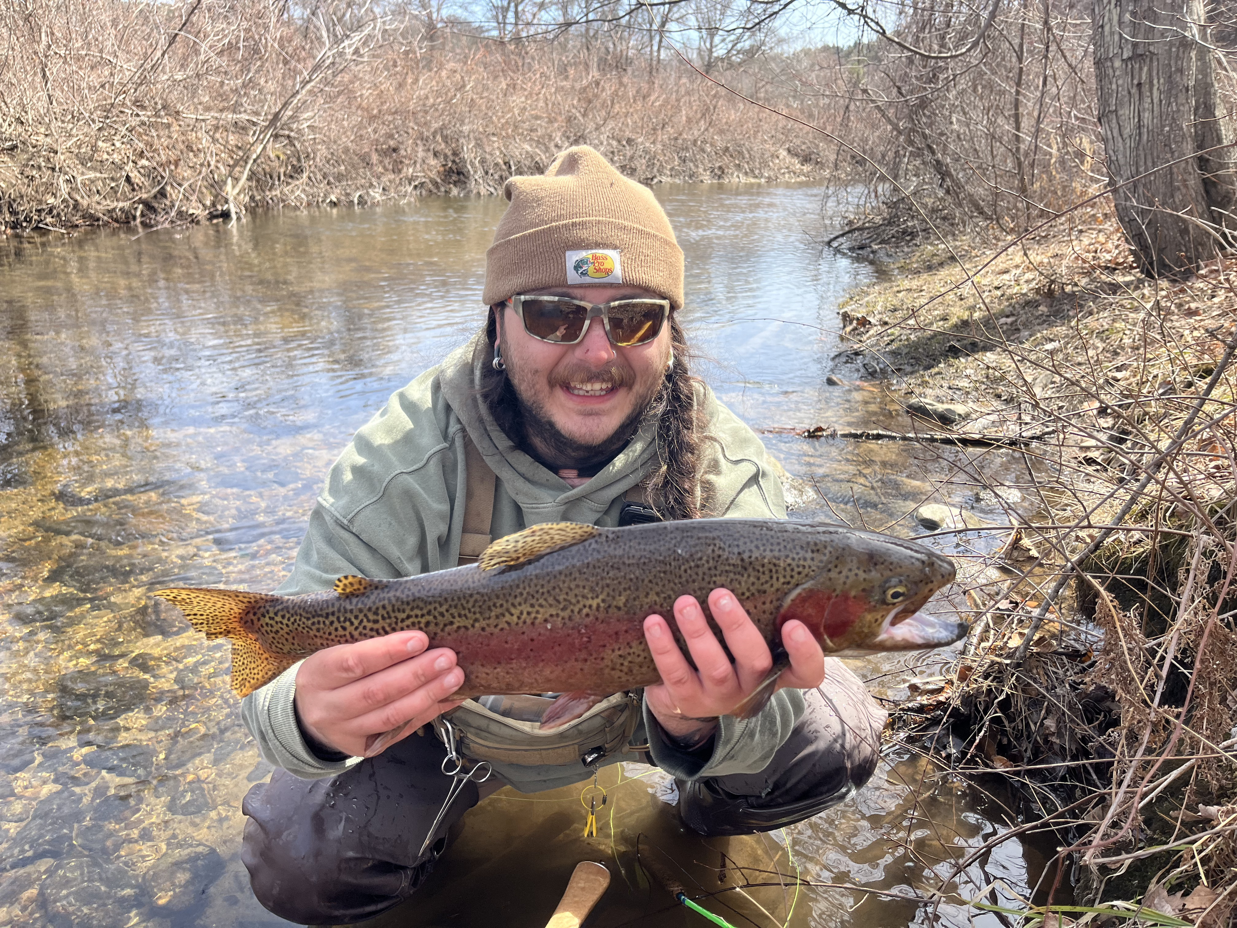 Man kneeling in shallow river water, holding a rainbow trout, wearing sunglasses, a tan beanie, and outdoor clothing, surrounded by leafless trees.